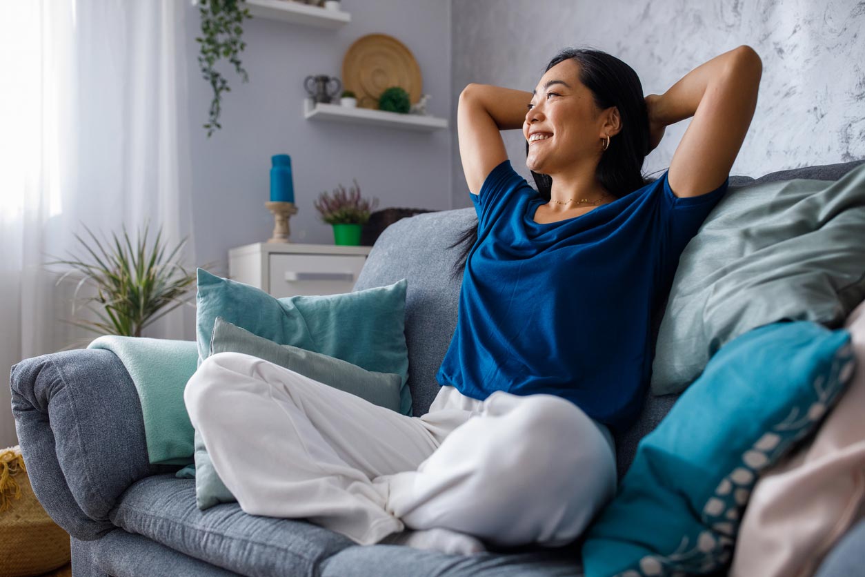 A woman sits comfortably on a sofa with colorful cushions, smiling and relaxing with her hands behind her head in a cozy, well-lit living room.