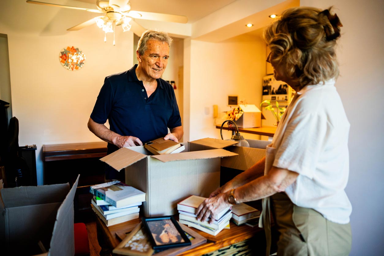 An older man and woman pack books into cardboard boxes in a well-lit dining room. The man is smiling, and the woman is stacking books. Framed photos and other items are on the table and furniture nearby.