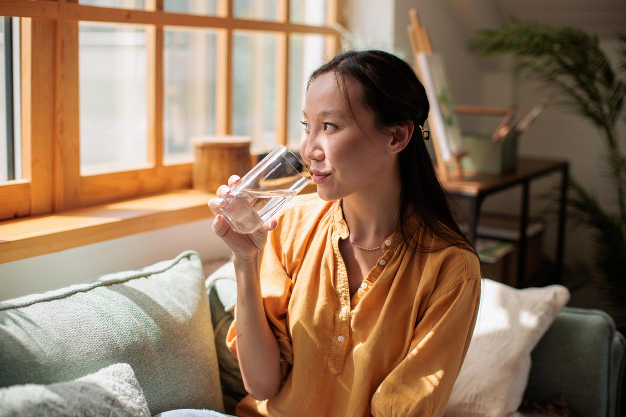 A woman in a yellow shirt sits on a sofa by a sunny window, holding and drinking a glass of water, looking thoughtfully outside.