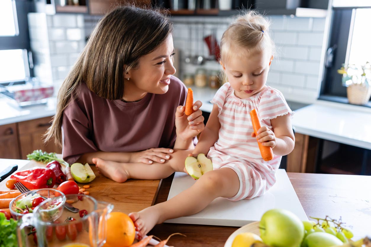 A woman smiles at a young girl sitting on a kitchen counter. Both are holding carrots. Fresh fruits and vegetables are spread on the counter around them, suggesting they are preparing a healthy meal together.