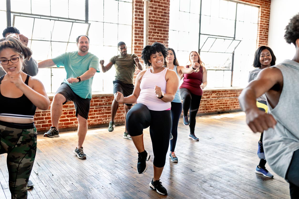 A diverse group of people in fitness attire participate in a lively dance exercise class in a spacious studio with large windows and brick walls, all smiling and following the instructor’s movements.