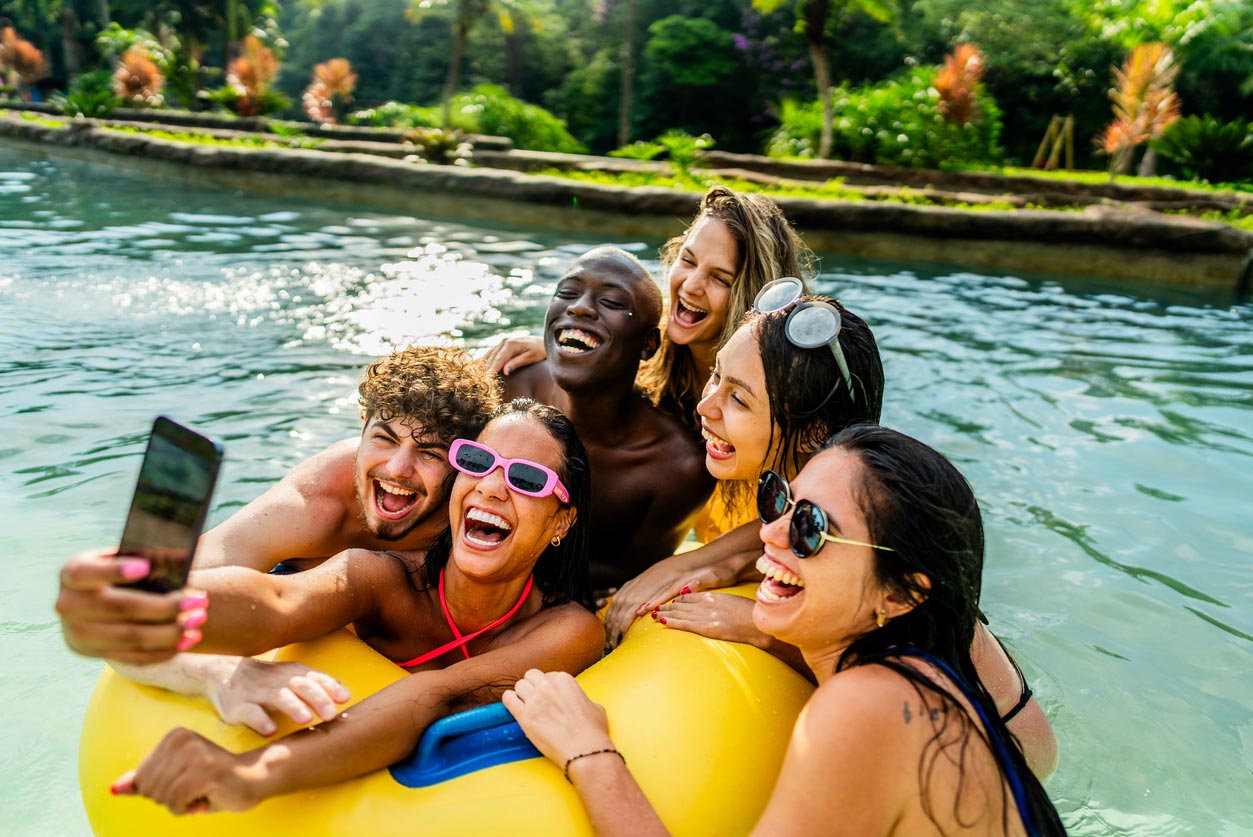 A group of five friends in swimwear smile and pose for a selfie on a yellow inflatable ring in a pool, surrounded by lush greenery and sunlight in the background.