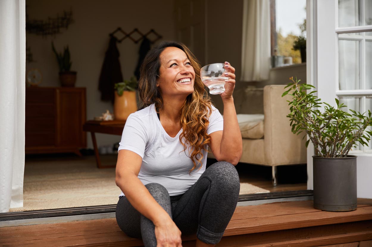 Smiling woman with long hair sits on wooden steps outside a house, holding a glass of water. She wears a white t-shirt and gray leggings, with plants and cozy furniture visible inside through open doors.