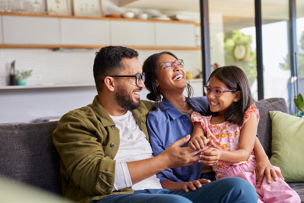 A man, woman, and young girl sit on a couch at home, laughing and enjoying each other’s company. They all wear glasses and casual clothes, and the background shows a modern, bright living room and kitchen.