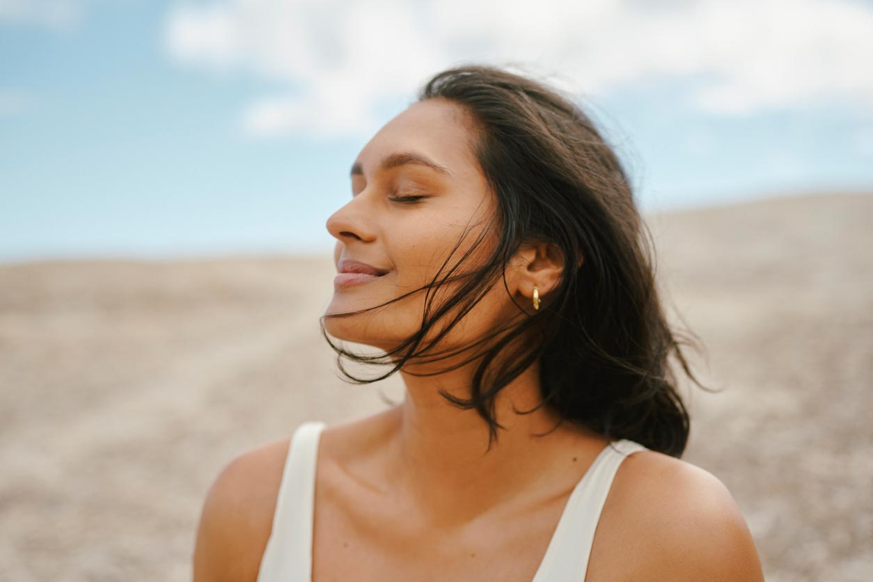 A woman with medium-length dark hair and gold hoop earrings smiles softly with her eyes closed, standing outdoors on a sunny day with a blue sky and blurred sandy background.