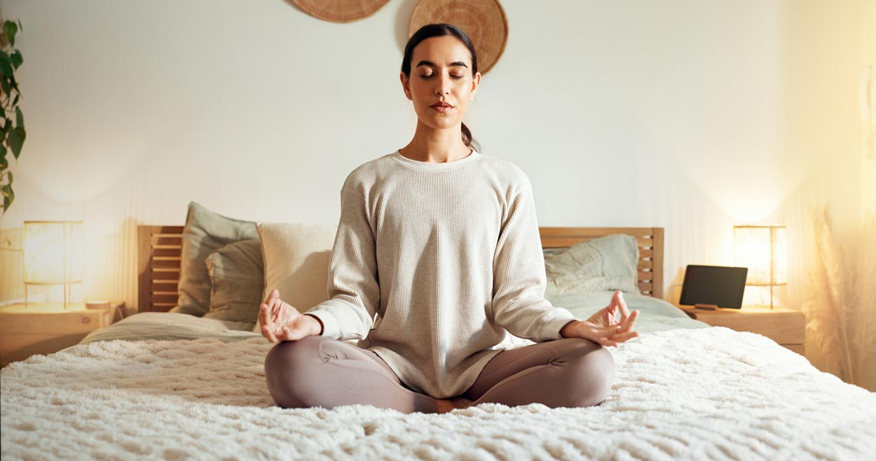 A woman sits cross-legged on a bed with eyes closed, meditating in a calm, softly lit bedroom with neutral decor and cozy bedding.