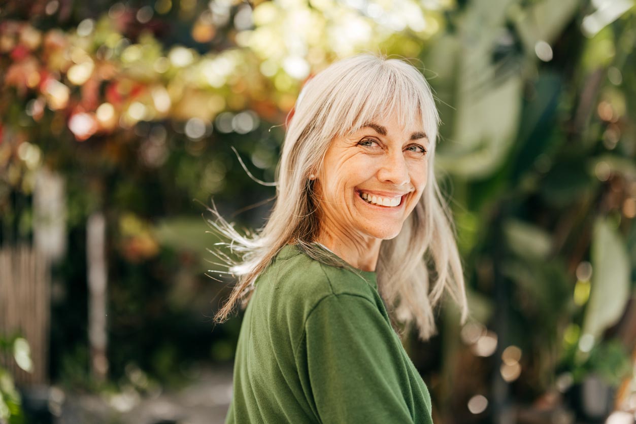 A smiling older woman with gray hair stands outdoors, wearing a green shirt. She looks over her shoulder with a cheerful expression, with blurred greenery and sunlight in the background.