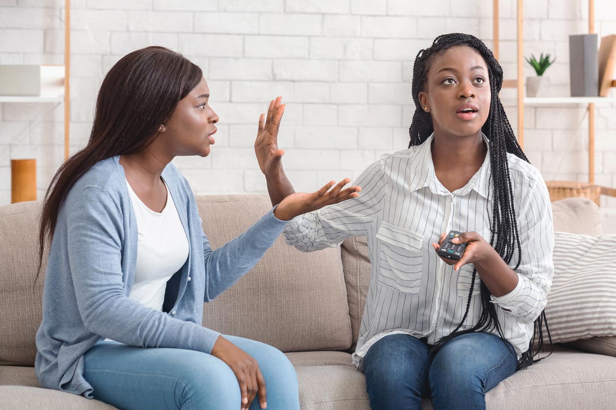 Two women sit on a couch. One, holding a remote, raises her hand as if to stop the other. The second woman gestures with frustration. Both appear to be having a disagreement.