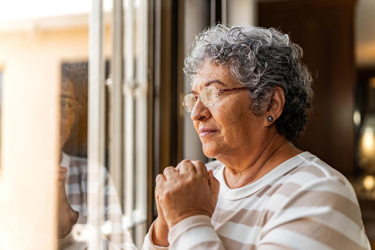 An elderly woman with gray hair and glasses stands by a window, hands clasped together, eyes closed, and appears peaceful and thoughtful. She is wearing a light-colored striped top.