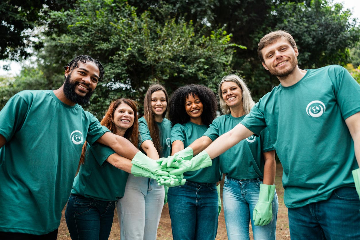 A diverse group of six people wearing green shirts and gloves stand outdoors, smiling and stacking their hands together in a gesture of teamwork and unity. Trees and greenery are visible in the background.