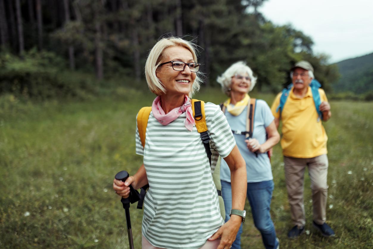 Three older adults with backpacks are hiking through a grassy area near a forest. One woman in front smiles, holding trekking poles, while the two others, a woman and a man, walk behind her on a nature trail.