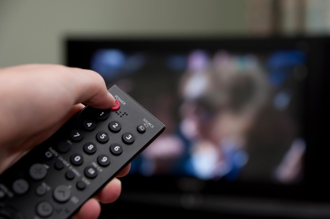 A person holds a remote control, pointing it at a blurred television screen in the background, suggesting they are about to turn on or change the channel.