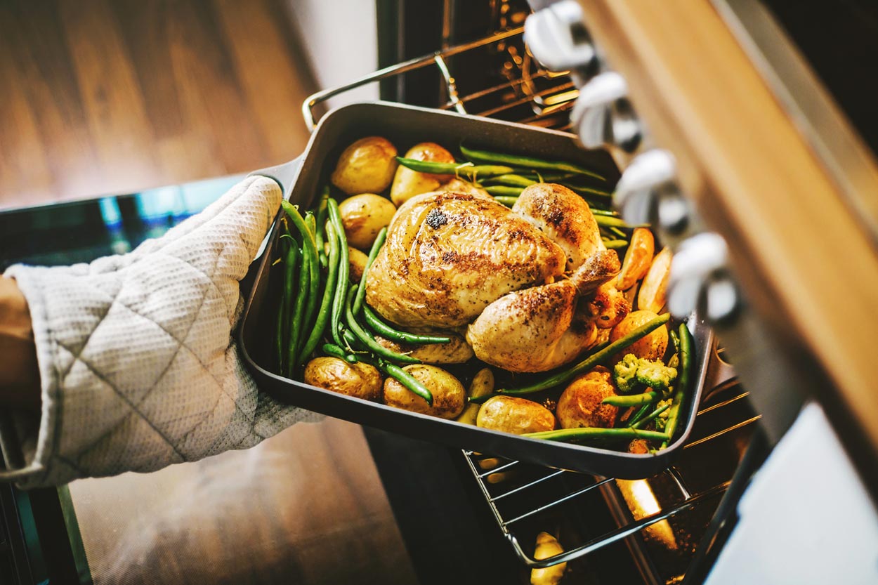 A person wearing an oven mitt removes a pan of roasted chicken with green beans, potatoes, and carrots from an oven. The food looks golden and freshly cooked.