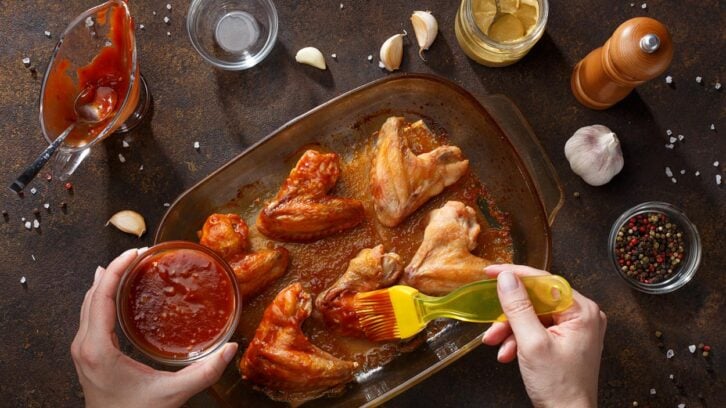 Overhead view of hands brushing barbecue sauce onto chicken wings in a glass baking dish, surrounded by garlic, salt, pepper, chips, and condiments on a dark table.