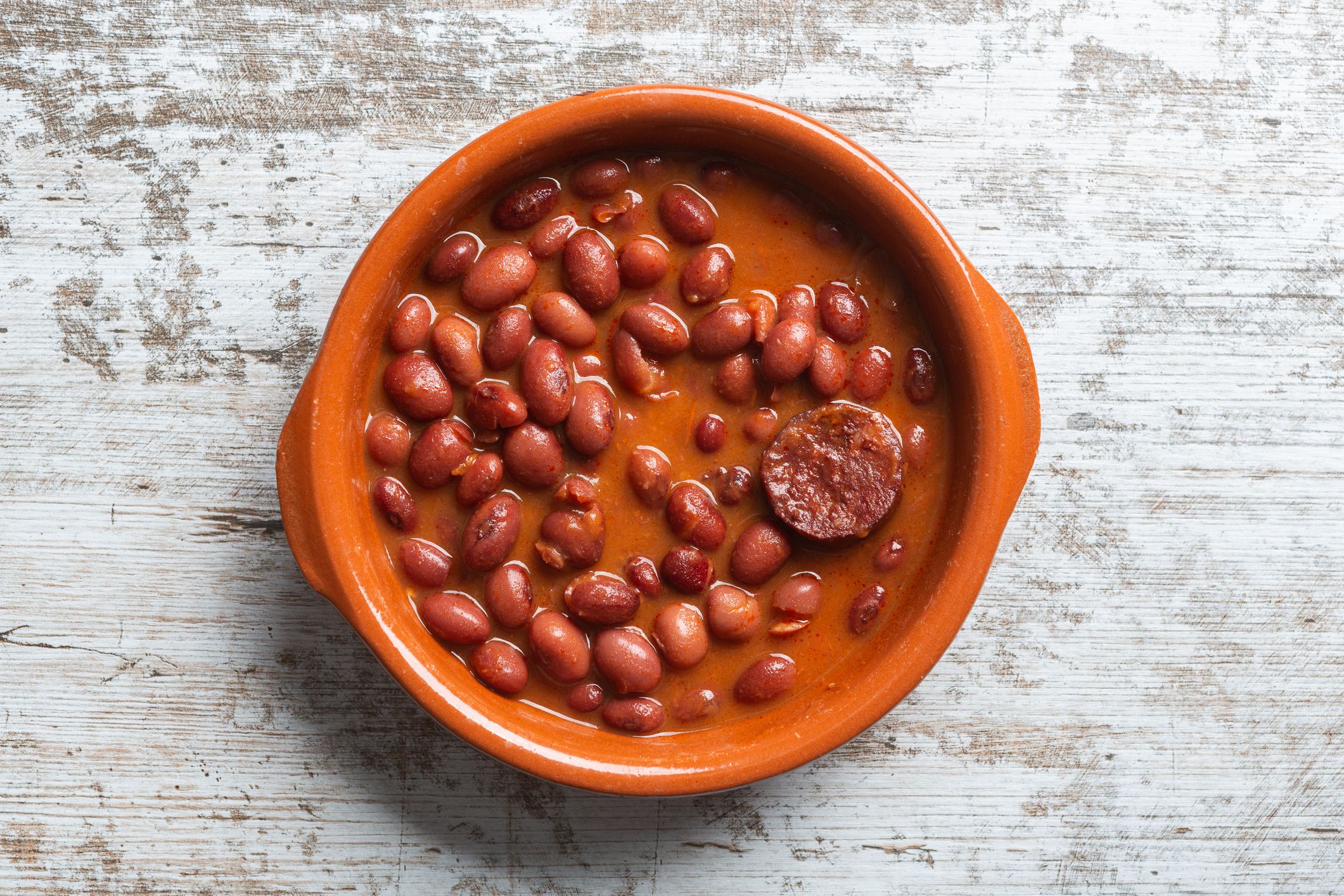 Dish of red beans in clay pot, on wooden table and red checkered tablecloth.
