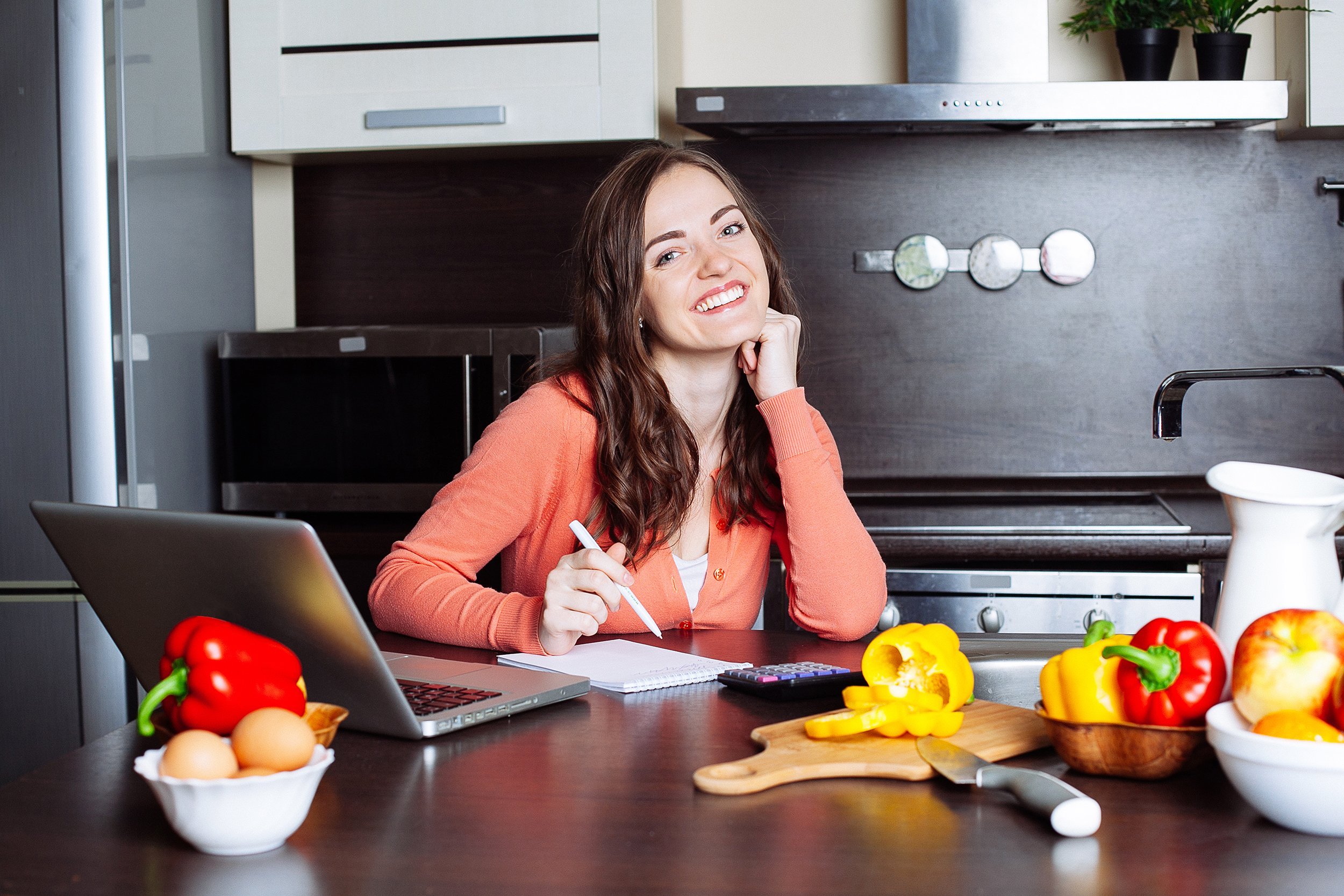 woman in her kitchen writing a grocery list with laptop, calculator, pen, and pencil