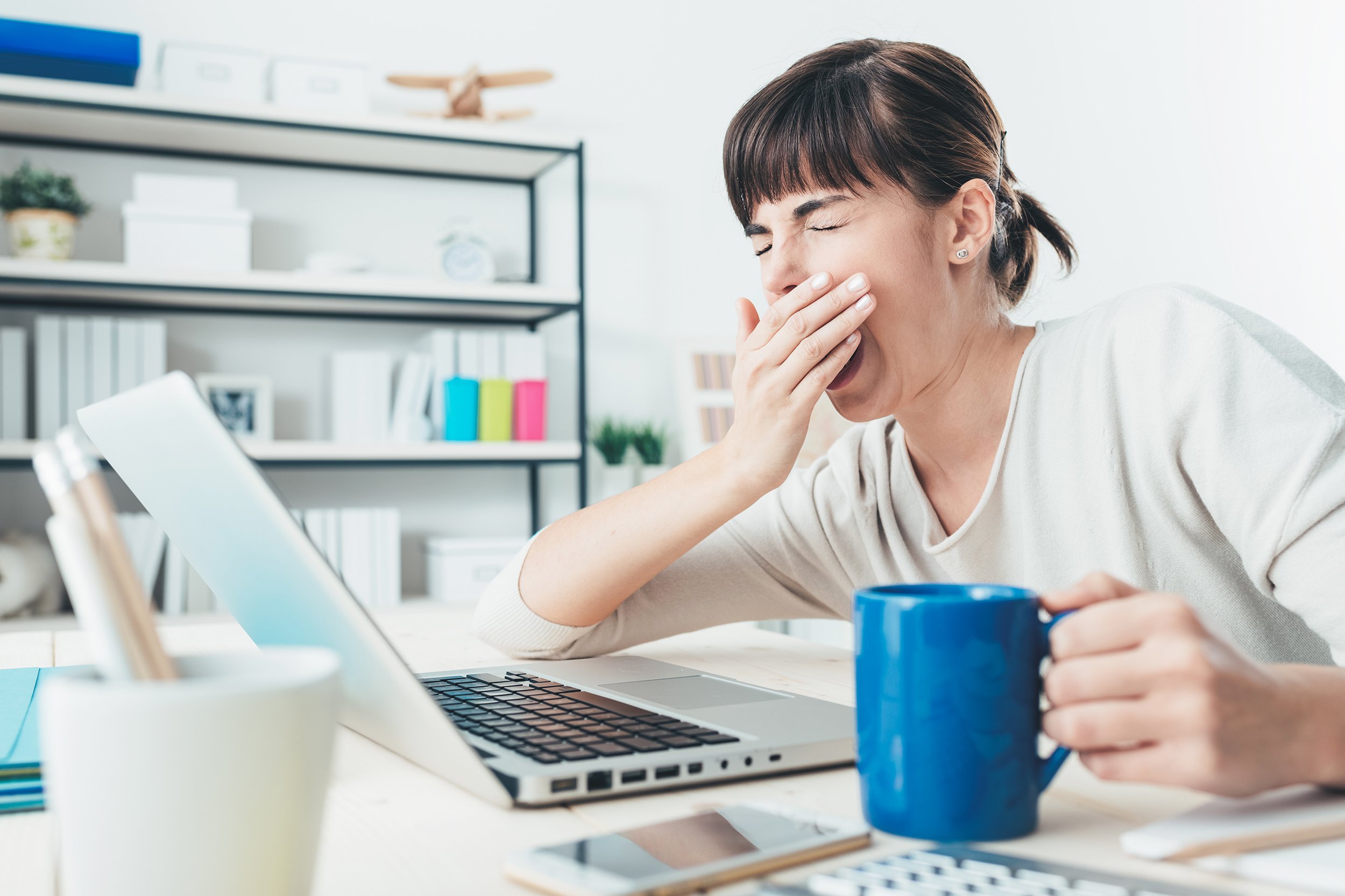 tired woman yawning working at office desk and holding a cup of coffee