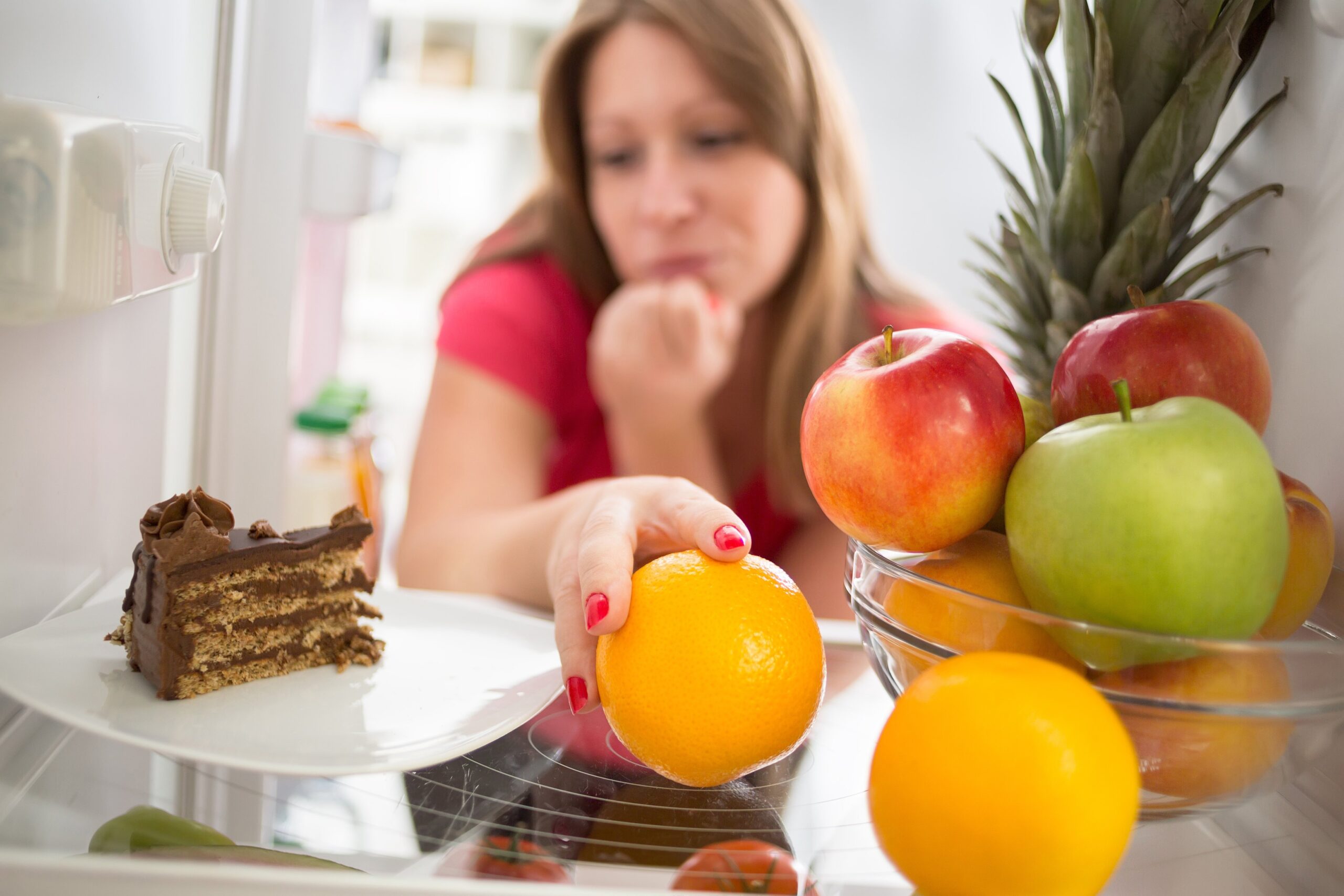 Woman reaches for orange in refrigerator while staring longingly at a piece of chocolate cake also within reach