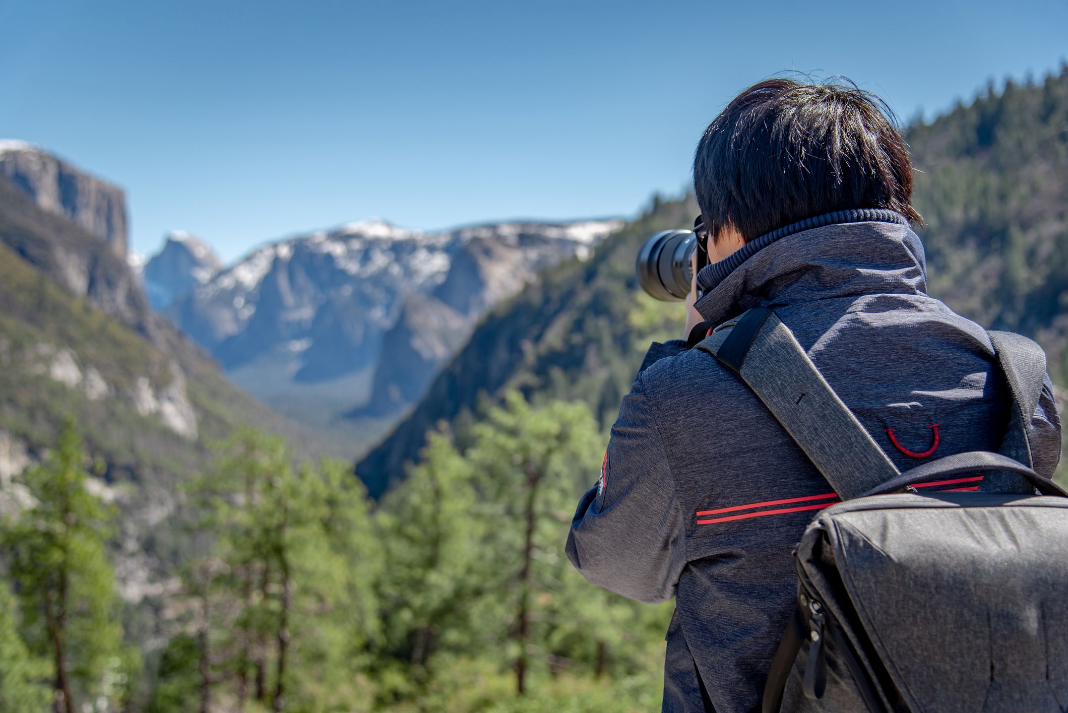 Asian man photographer and tourist with backpack holding DSLR camera taking photo of Half Dome mountain in Yosemite National Park, California.