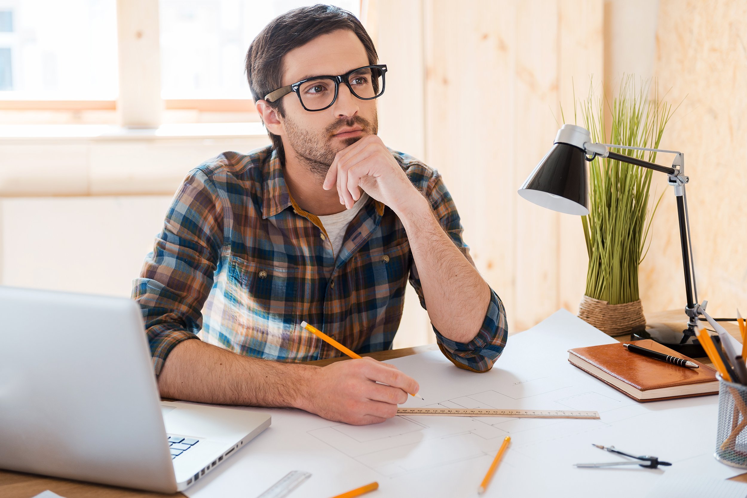 thoughtful young man holding hand on chin and looking away while sitting at his working place