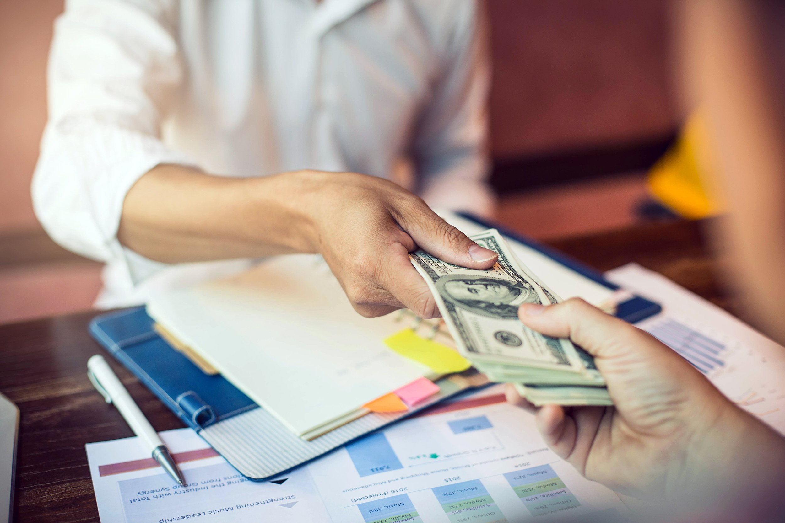 hands giving batch of hundred dollar bills to other hand over paperwork