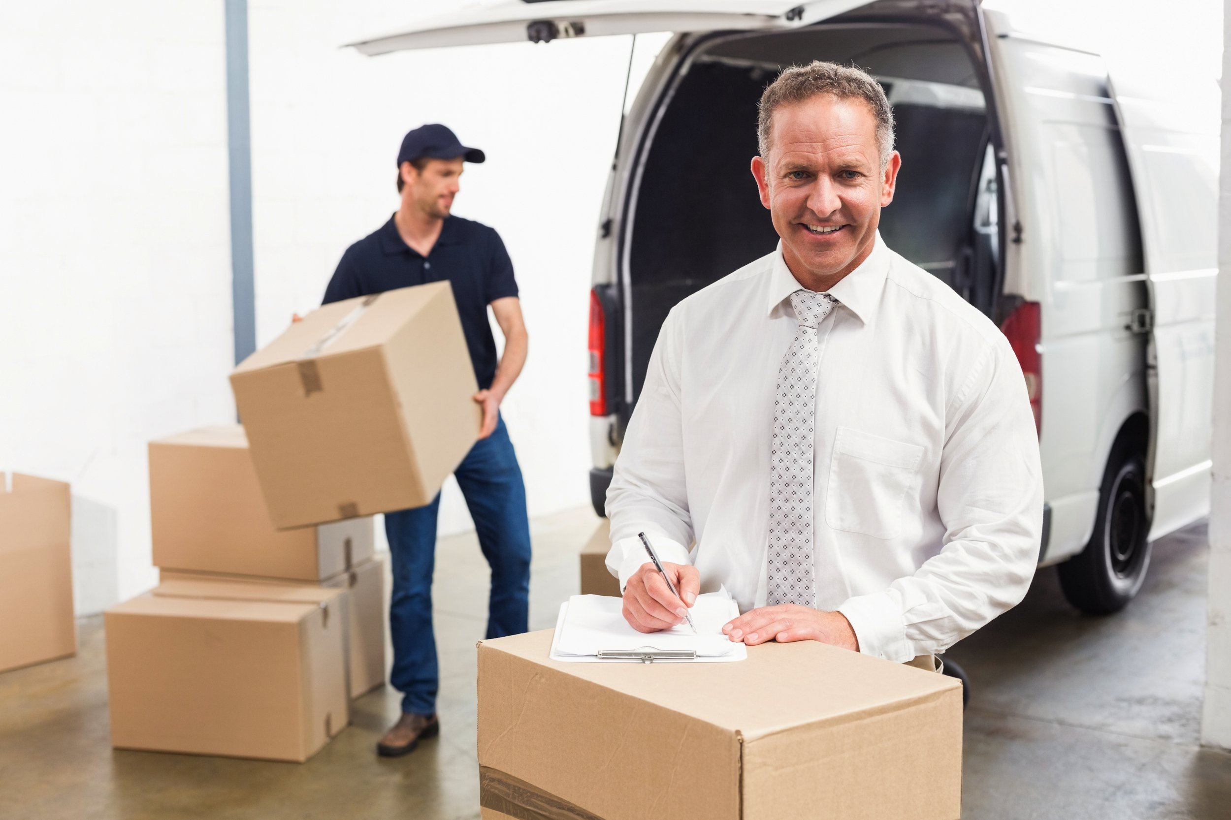 Smiling manager standing behind stack of cardboard boxes in a large warehouse.