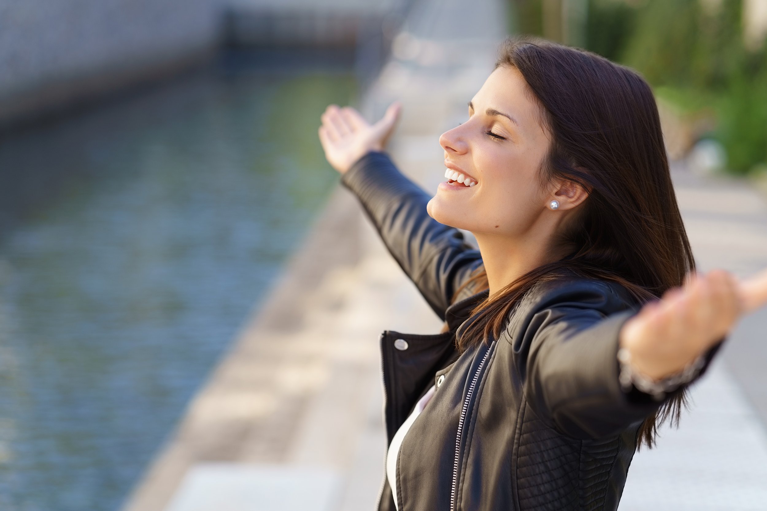 grateful woman with eyes closed and arms extended standing near stone walkway and small canal