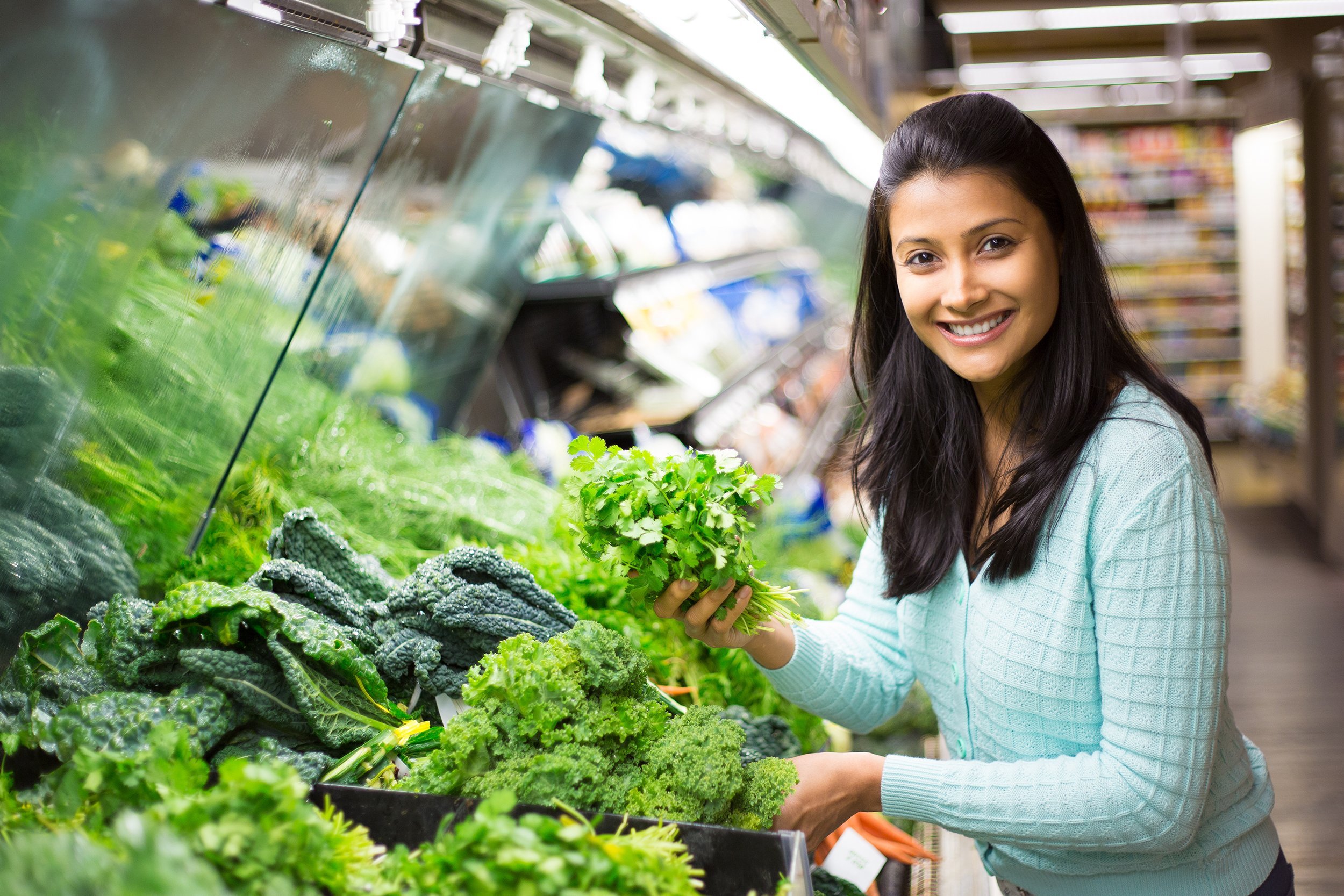 woman in sweater picking up green leafy vegetables in grocery store