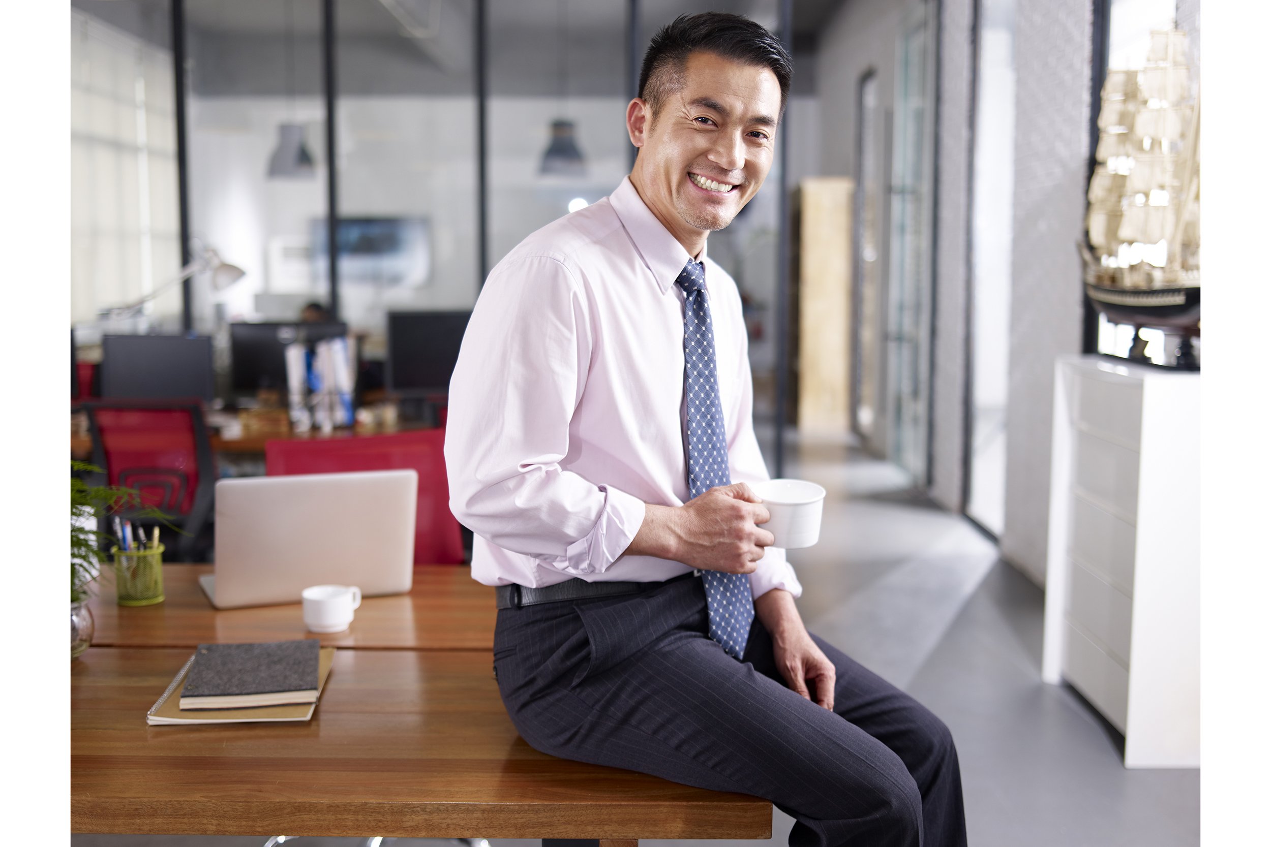 Asian businessman sitting on his desk smiling with a cup of coffee