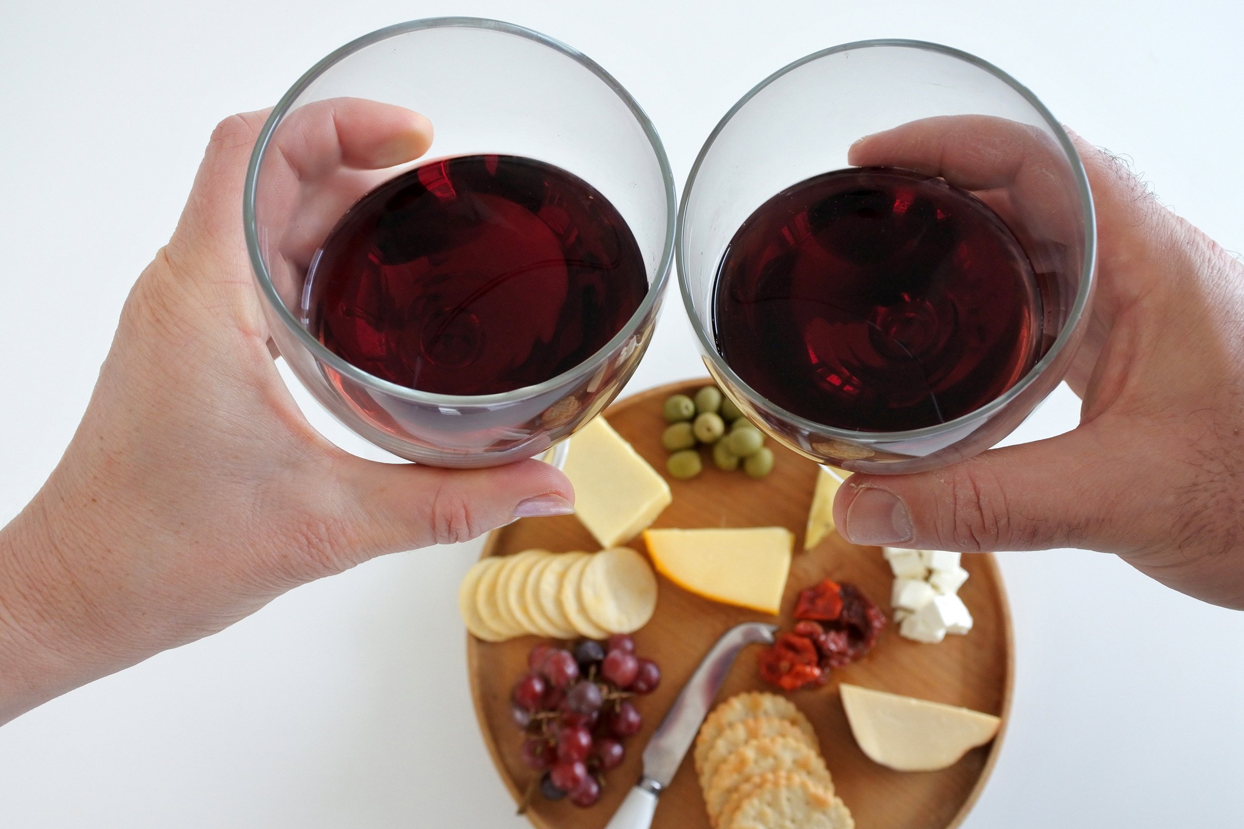 two hands holding glasses of red wine over cheese plate