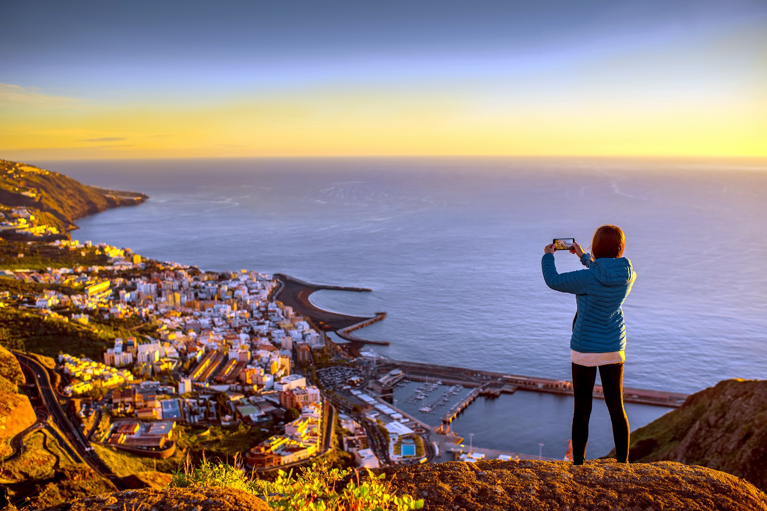 Woman standing on cliff taking photo of city below at sunset or sunrise
