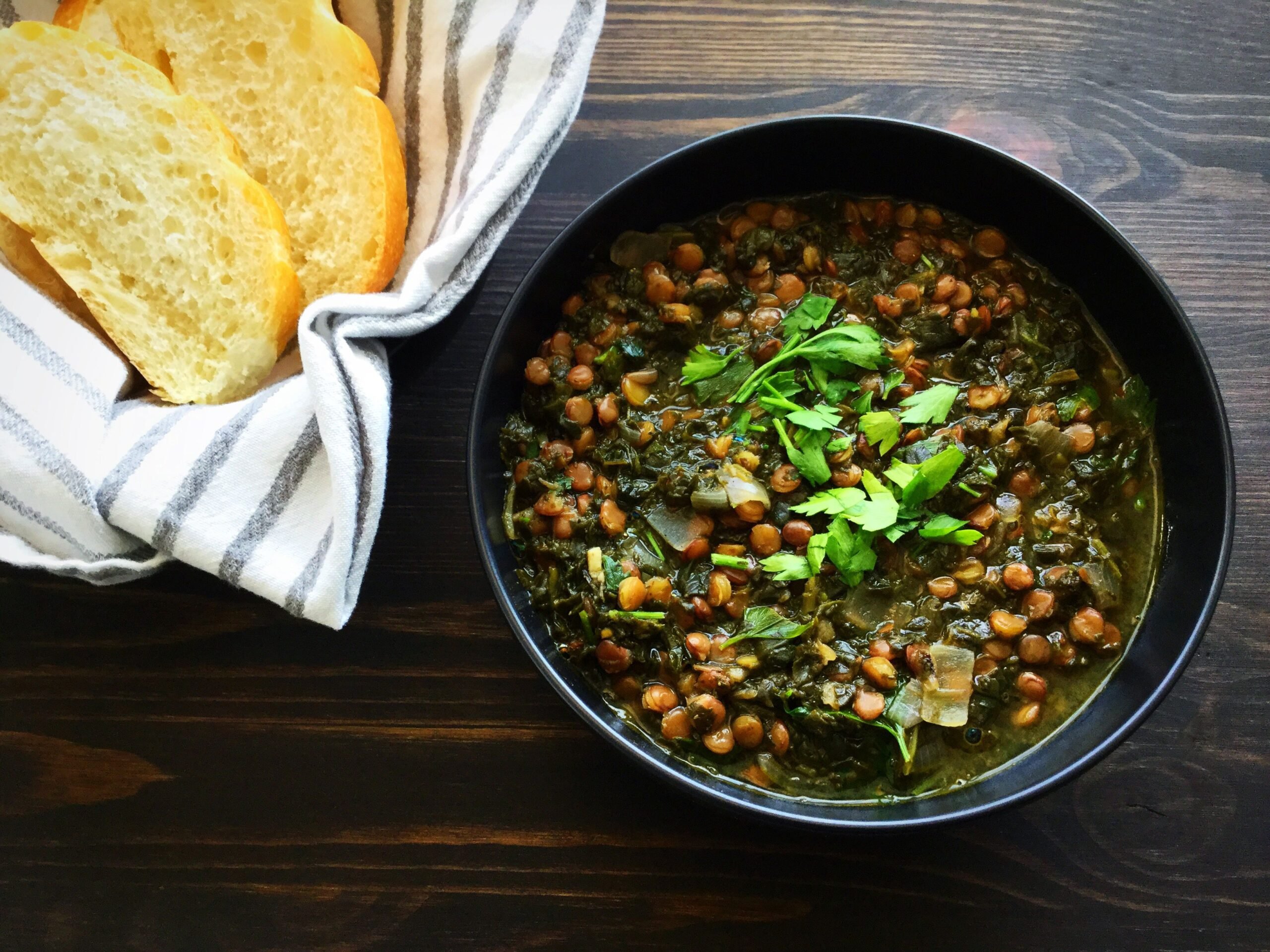 A bowl of spinach and lentil soup with slices of bread on the side shown on a dark wood background