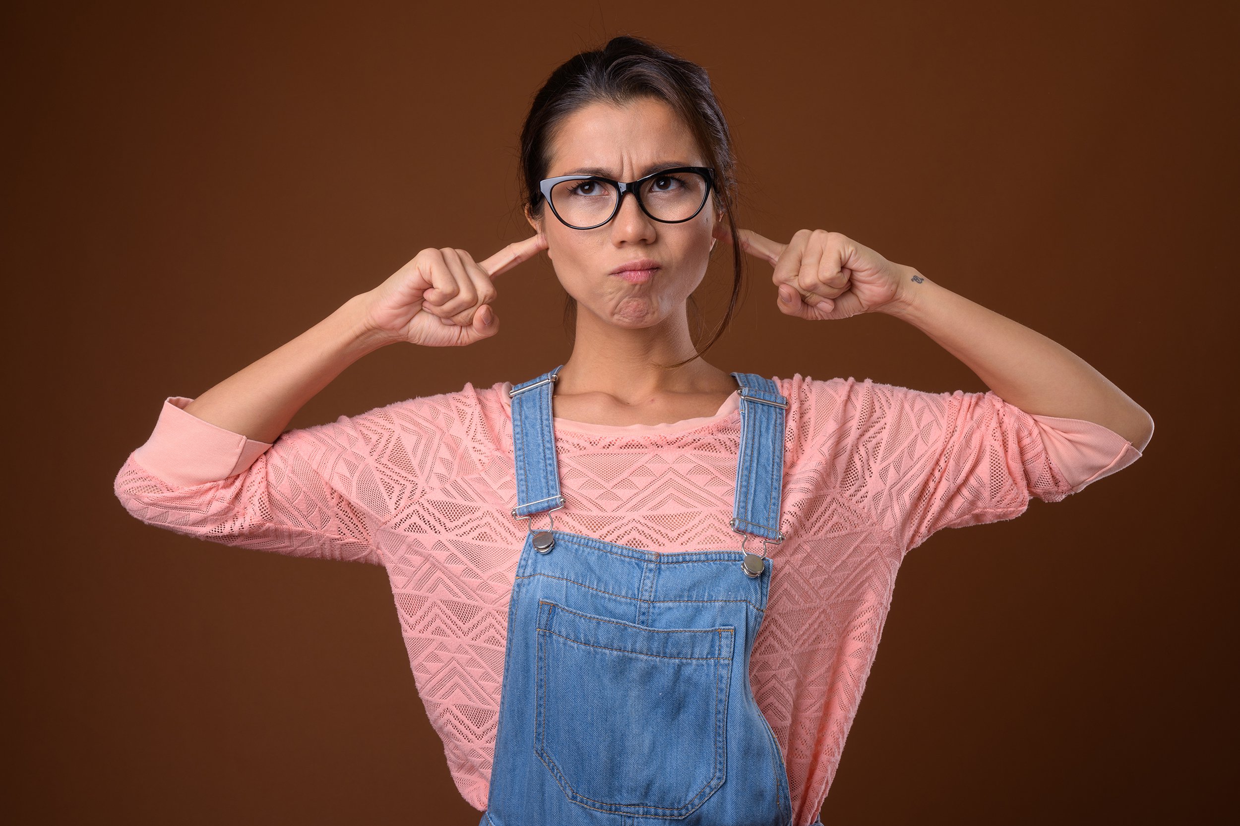 multi-ethnic woman wearing eyeglasses against colored background putting fingers in ears