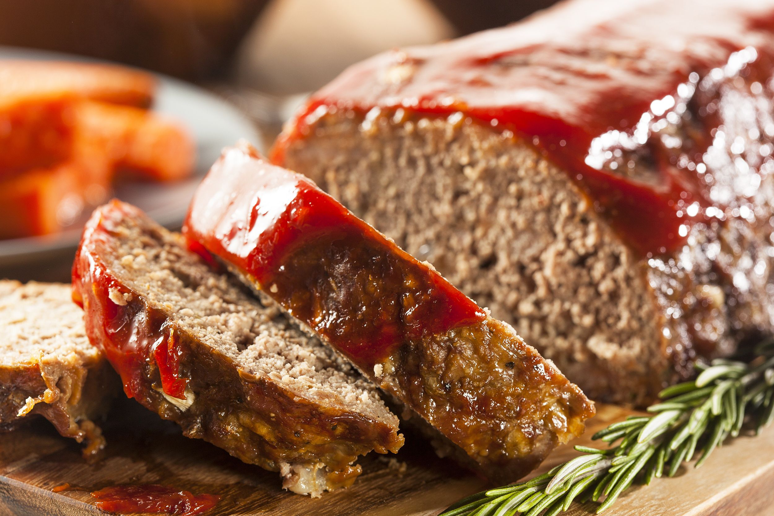 Closeup of two slices and whole meat loaf with herbs on a wooden cutting board with a blurred background of foods being served for a dinner