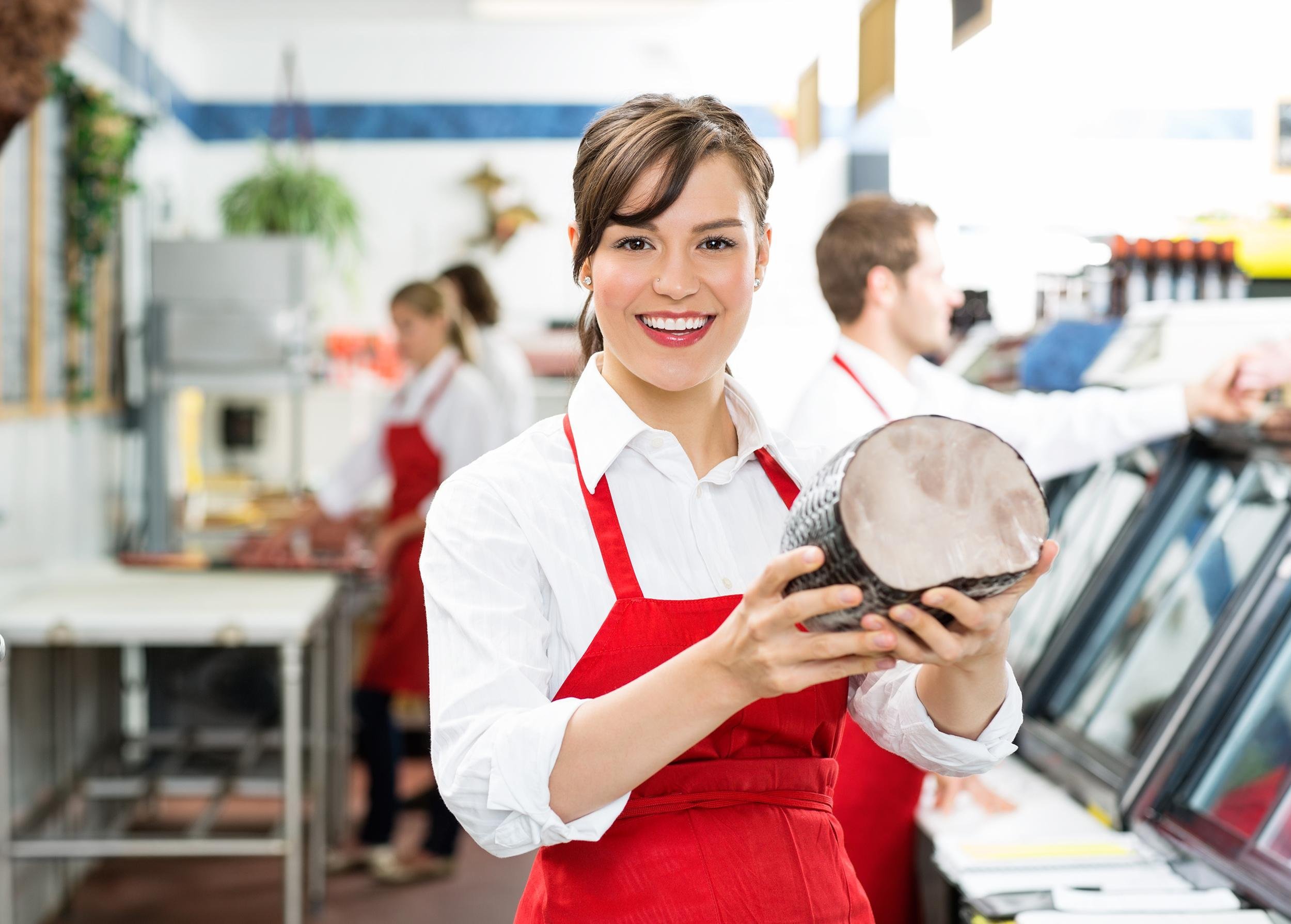 happy female butcher holding ham at store with colleagues working in background