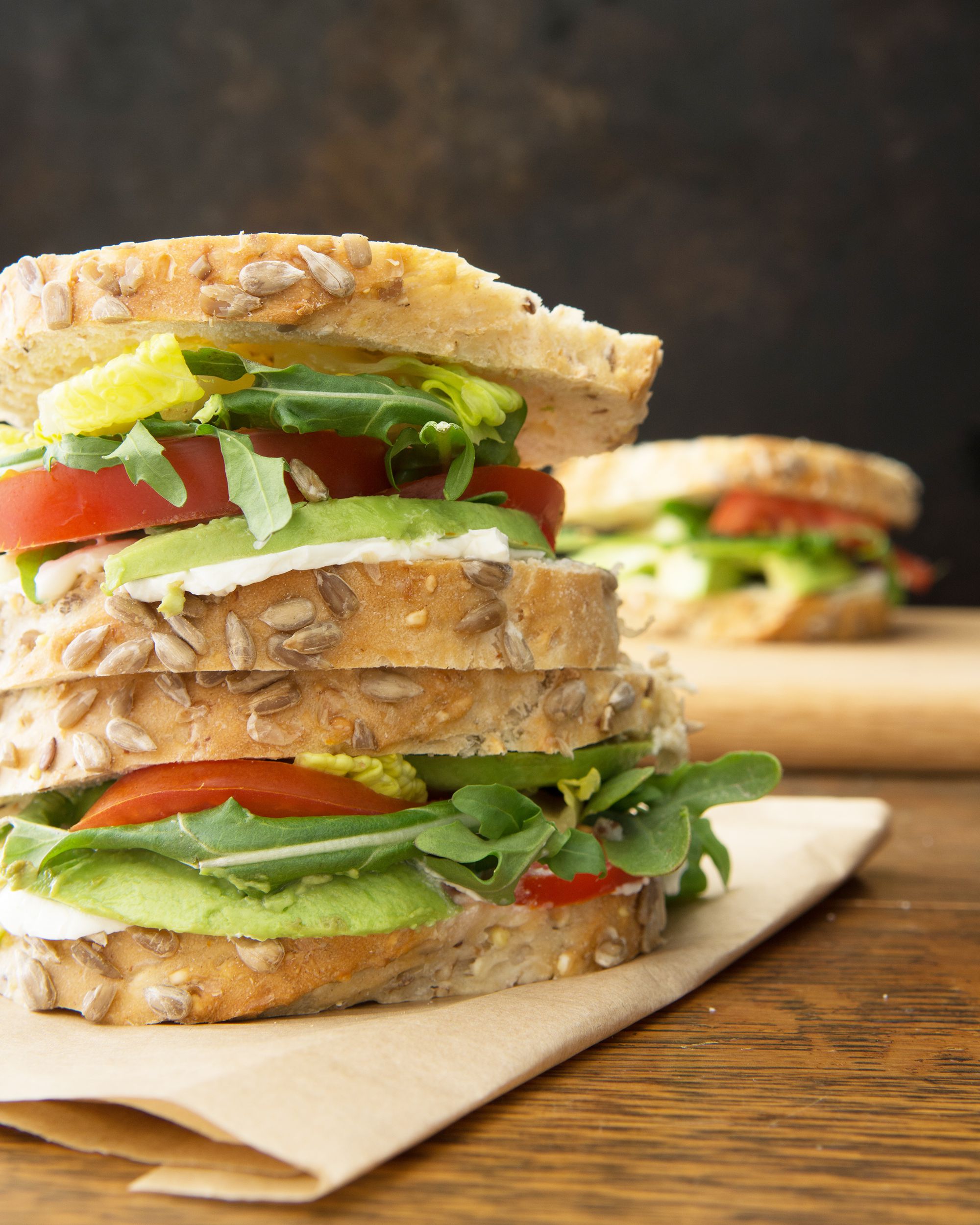 Closeup of two avocado, lettuce, and tomato sandwiches on top of each other on a brown napkin on a wooden table with another avocado, lettuce, and tomato sandwich blurred in the distance, a black background