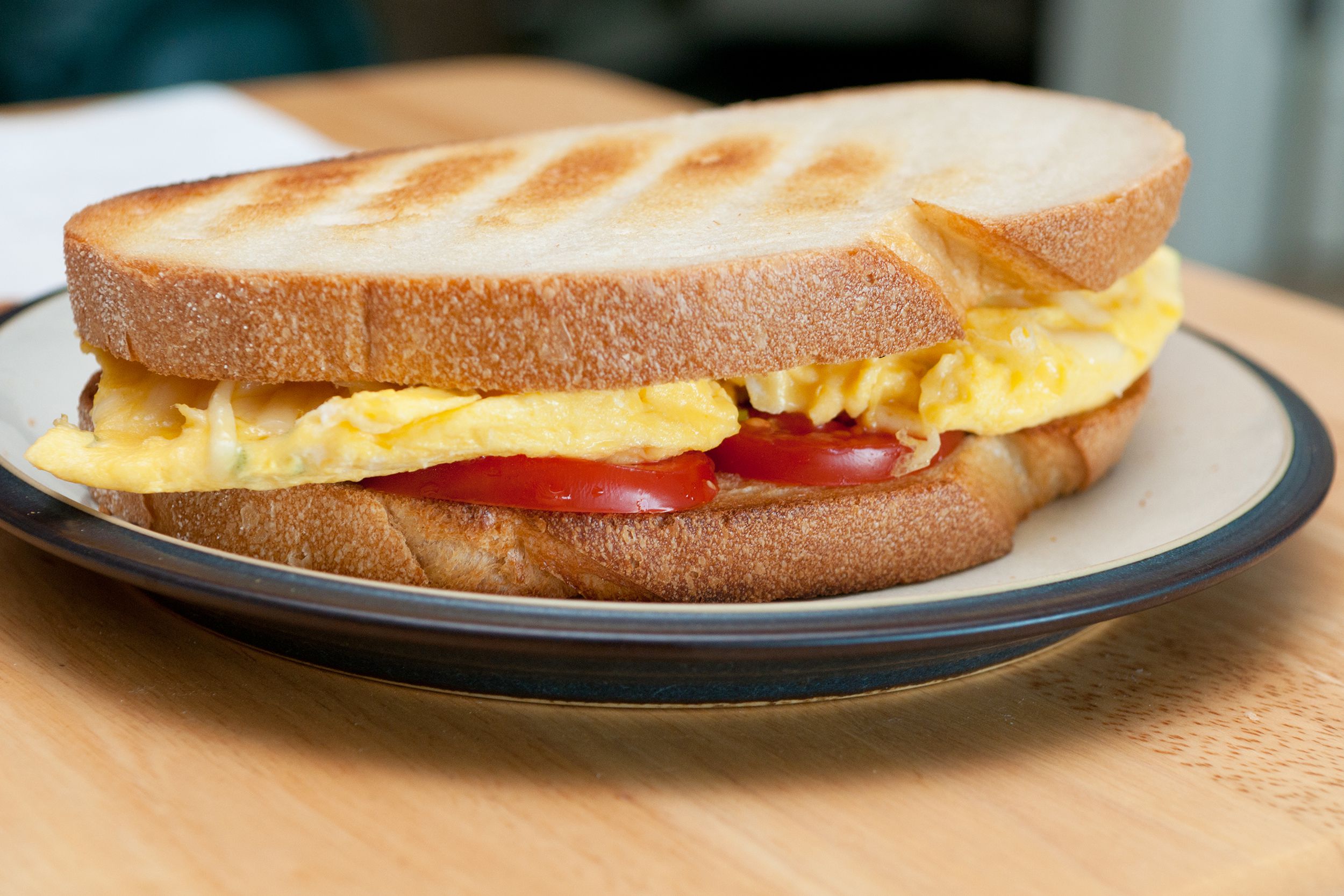 Egg sandwich with tomato on a brown-rimmed white ceramic plate, selective focus, on a wooden table with edge of table blurred in the background