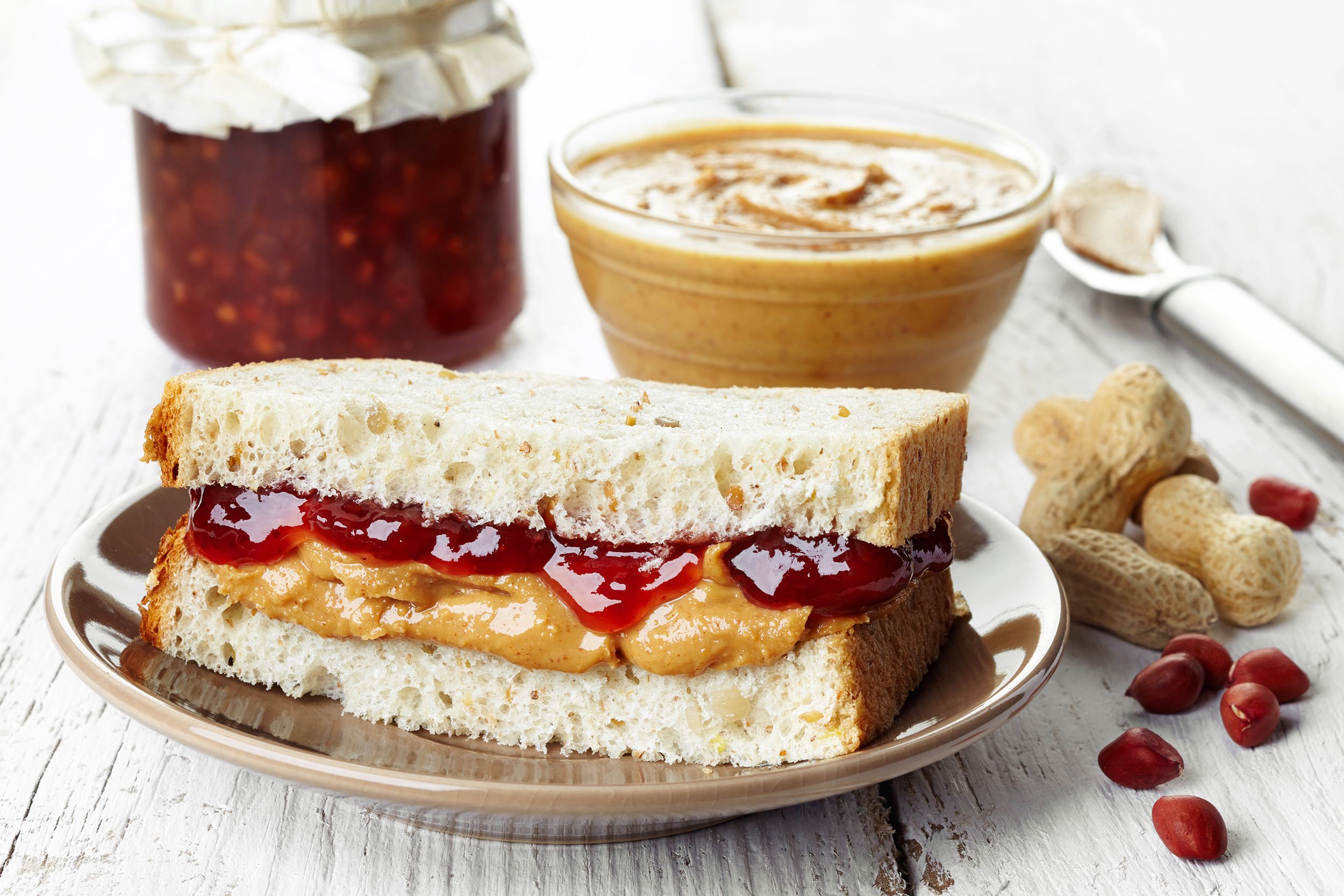 One half of peanut butter and jelly sandwich on a dark beige ceramic plate, a jar of strawberry jelly and a small glass bowl of peanut butter behind it on the top, on a rustic white wooden table, blurred in the background