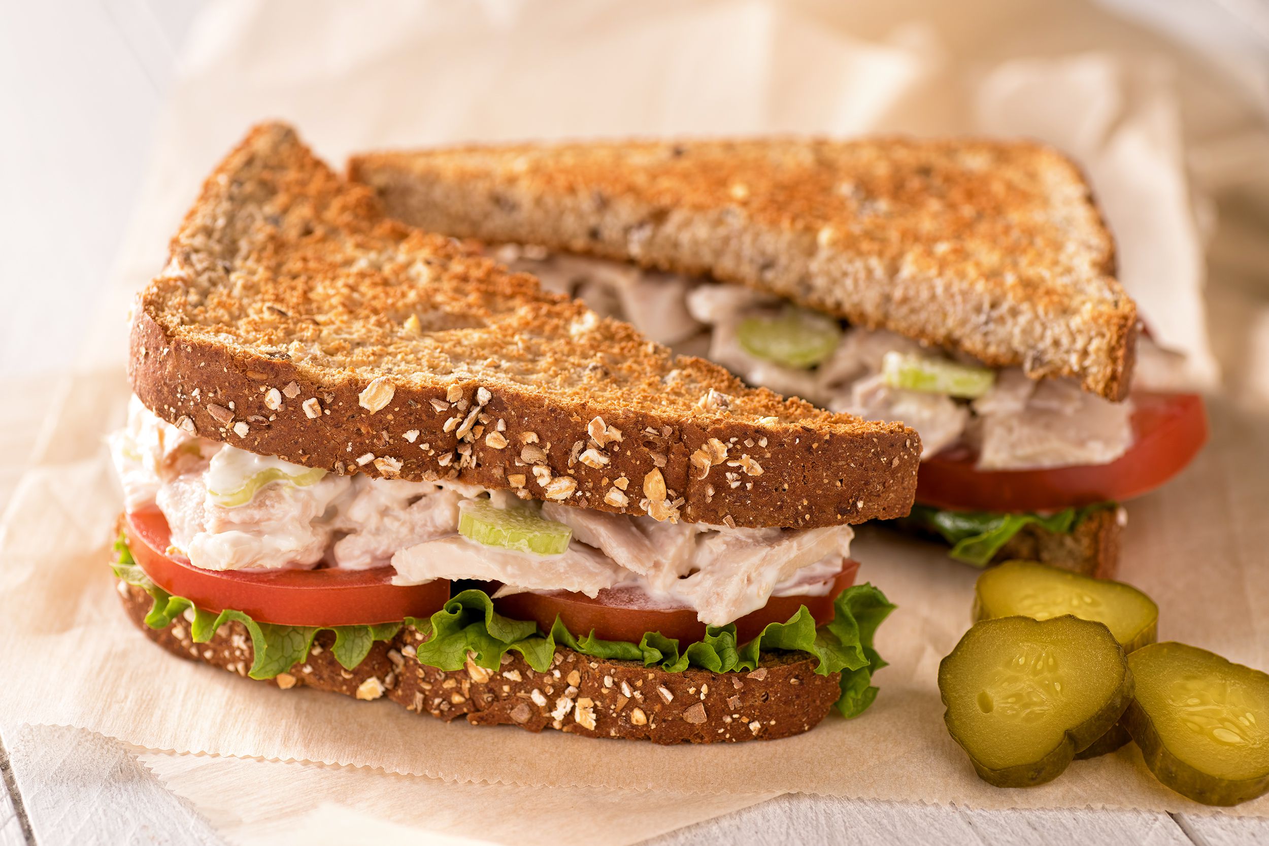 One tuna salad sandwich cut in half on a brown paper napkin with sliced pickles on a white wooden table