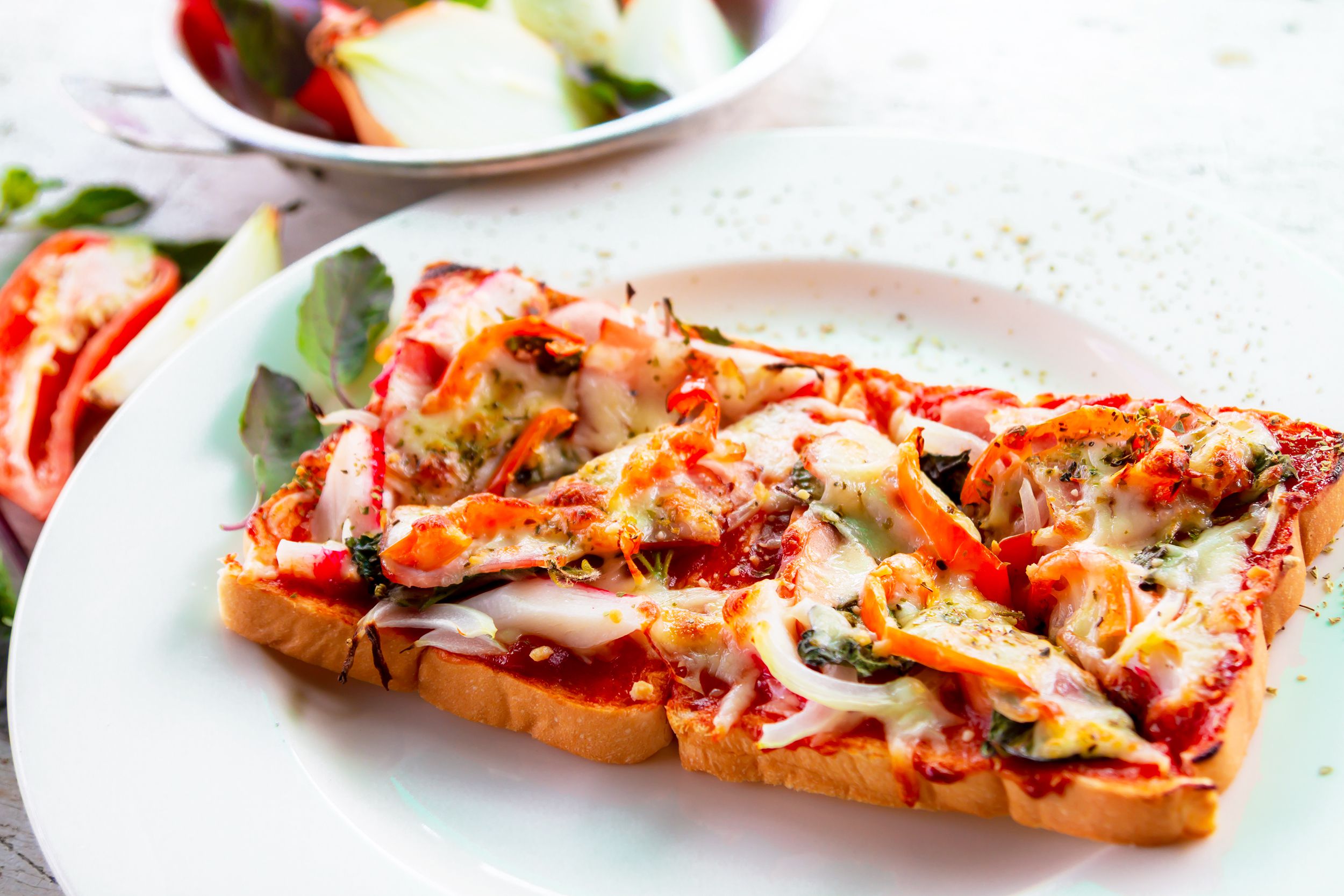 Pizza sandwich on a white porcelain plate, selective focus, herb sprinkled on the plate, surrounded by ingredients and a stainless steel bowl of a salad, blurred background of a white table in the top