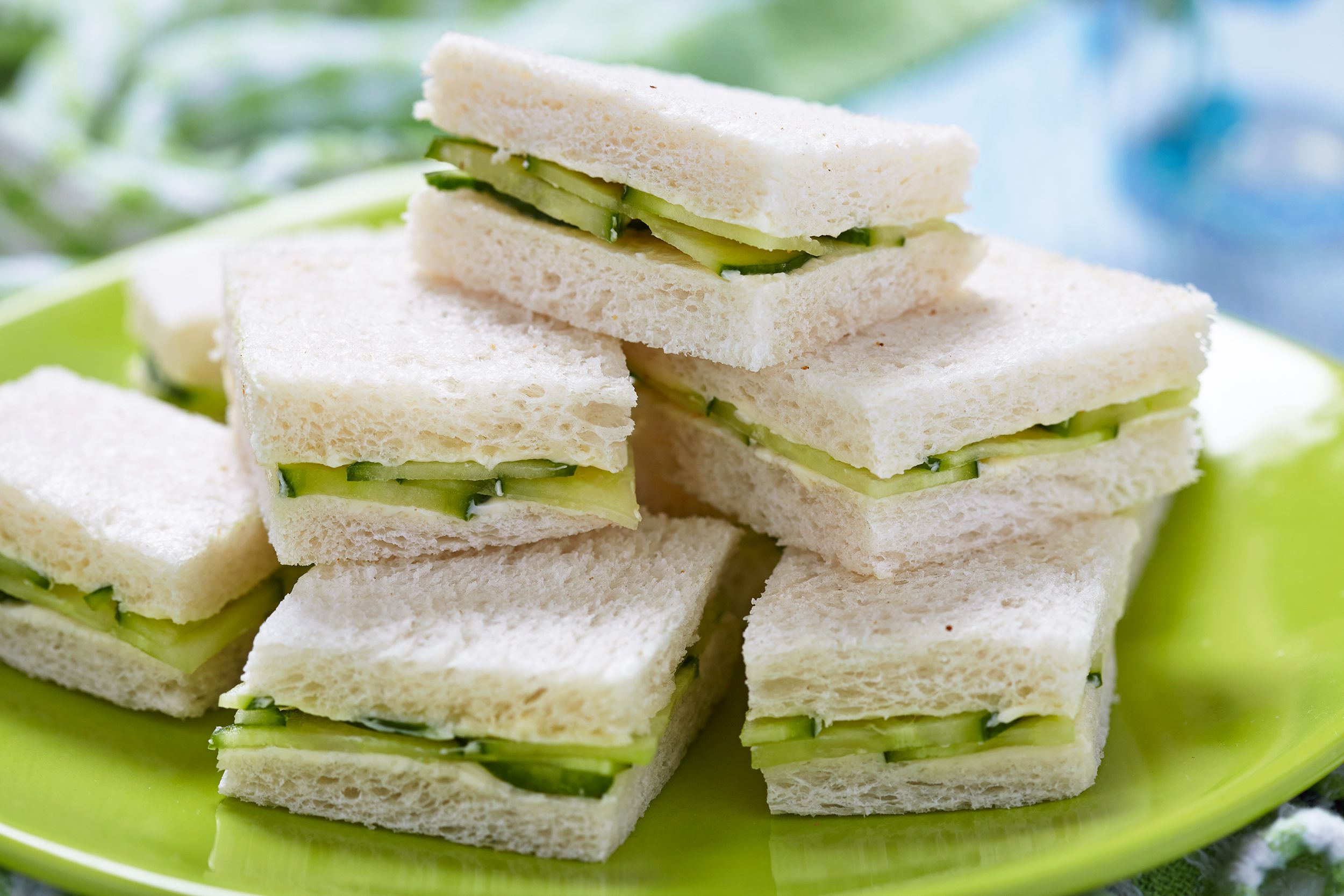 Several cucumber sandwich pieces piled as a pyramid on an avocado green ceramic plate, selective focus, an avocado green checkered napkin on the left and a light blue table on the right, blurred in the background