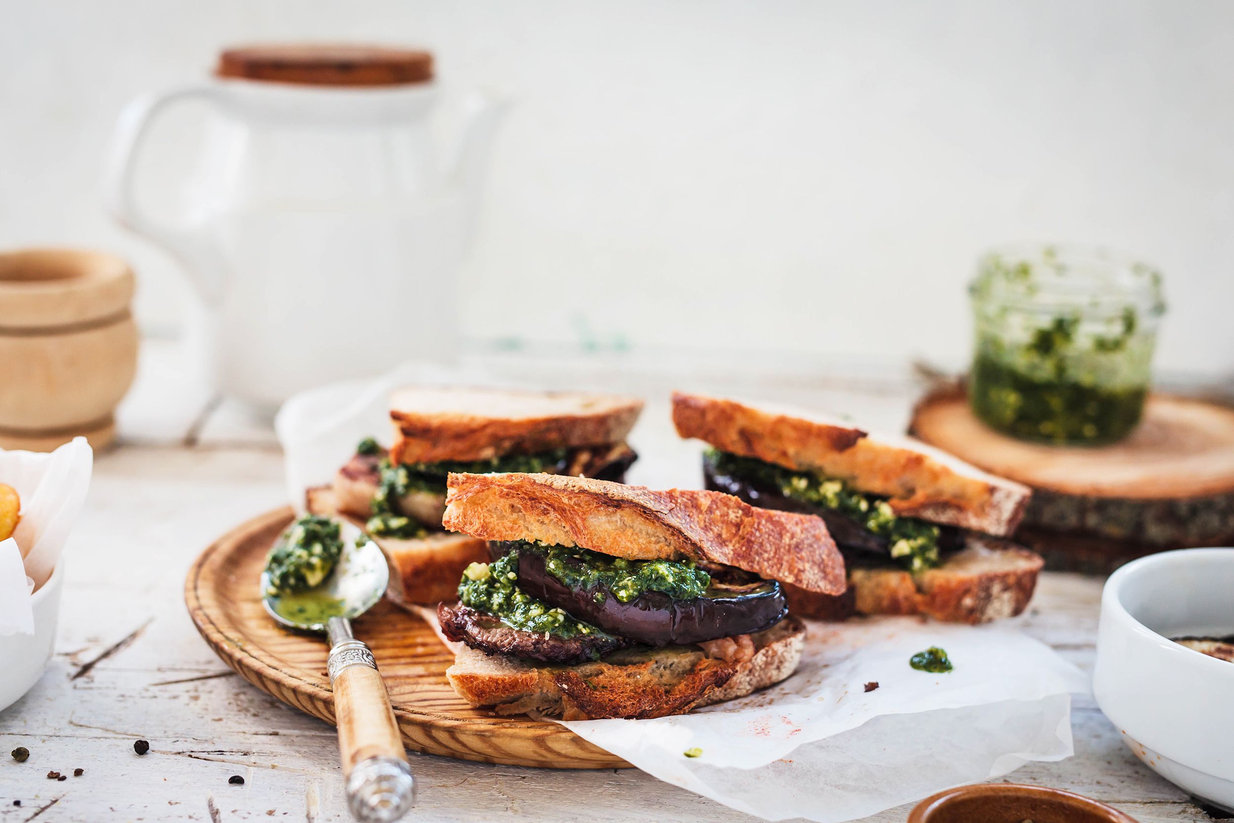 Three halves of balsamic roasted veggie sandwich on a rustic serving board with a spoon on the left with sauce, surrounded by ingredients, on a rustic white wooden table