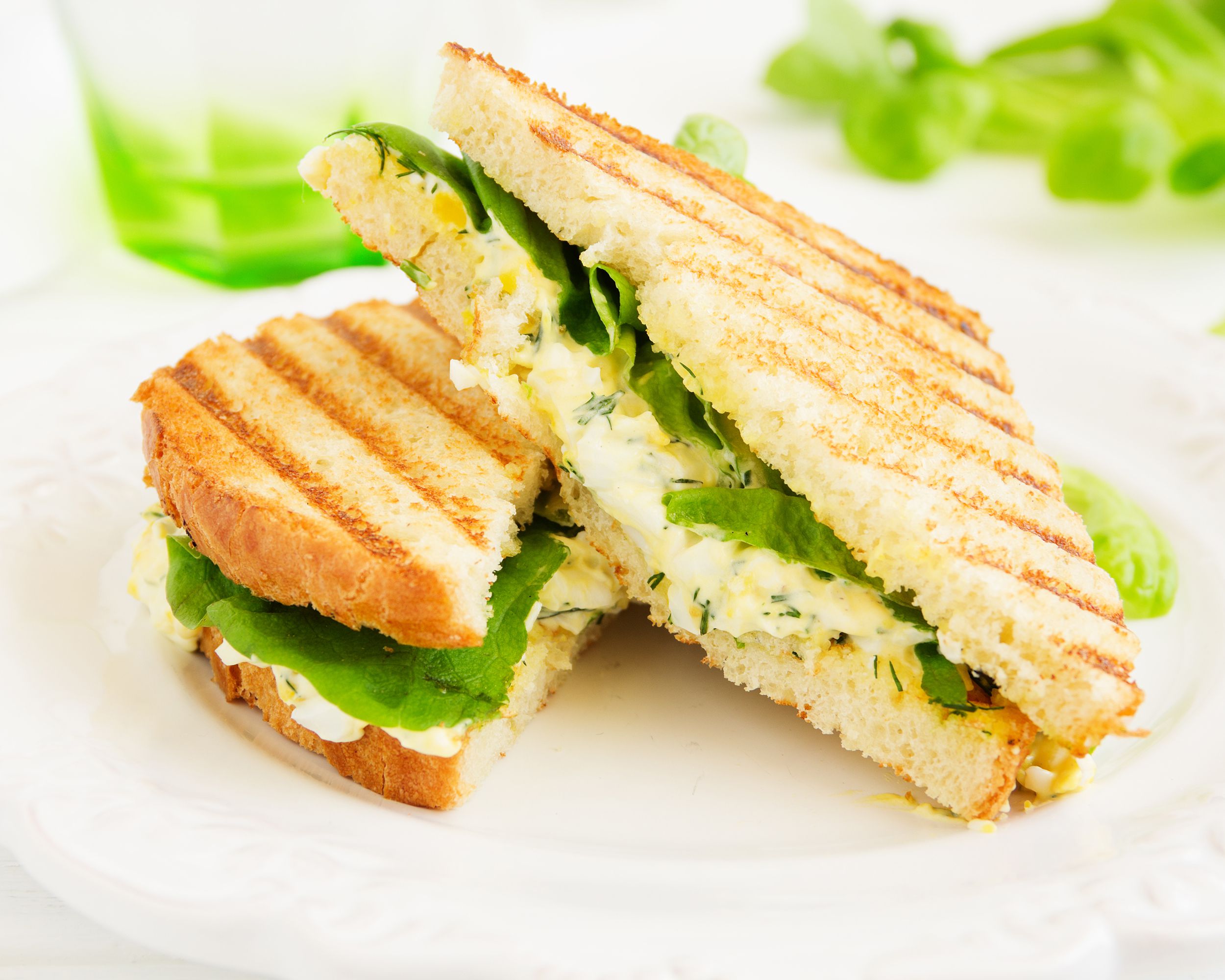 Two halves of egg salad sandwich piled on a fancy white porcelain plate, selective focus, on a white tablecloth, a green and clear glass and herb leaves blurred in the background