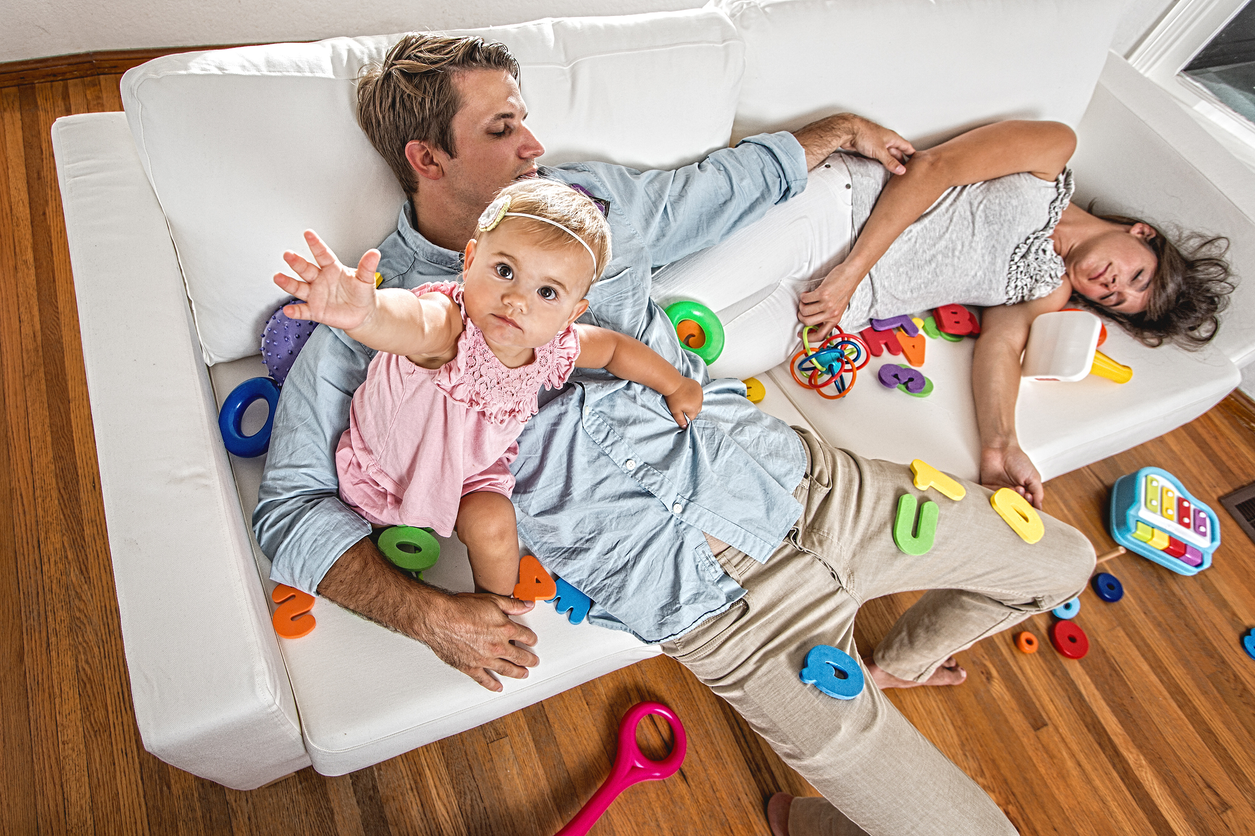 Exhausted parents asleep on couch next to awake baby and surrounded by toys