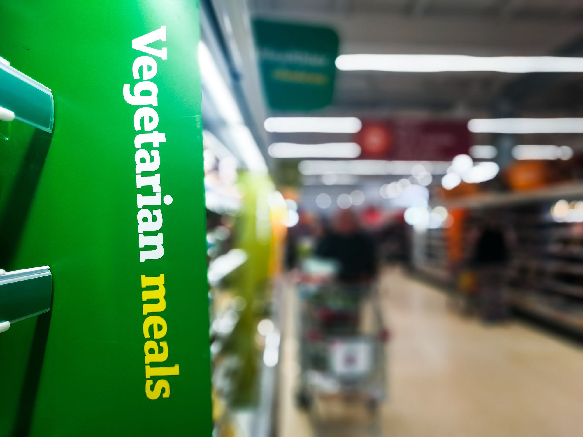 Close up color image depicting a Vegetarian Meals sign indicating the vegetarian food section inside a supermarket. Focus is on the sign, while the aisle of the supermarket is entirely defocused, allowing room for copy space.