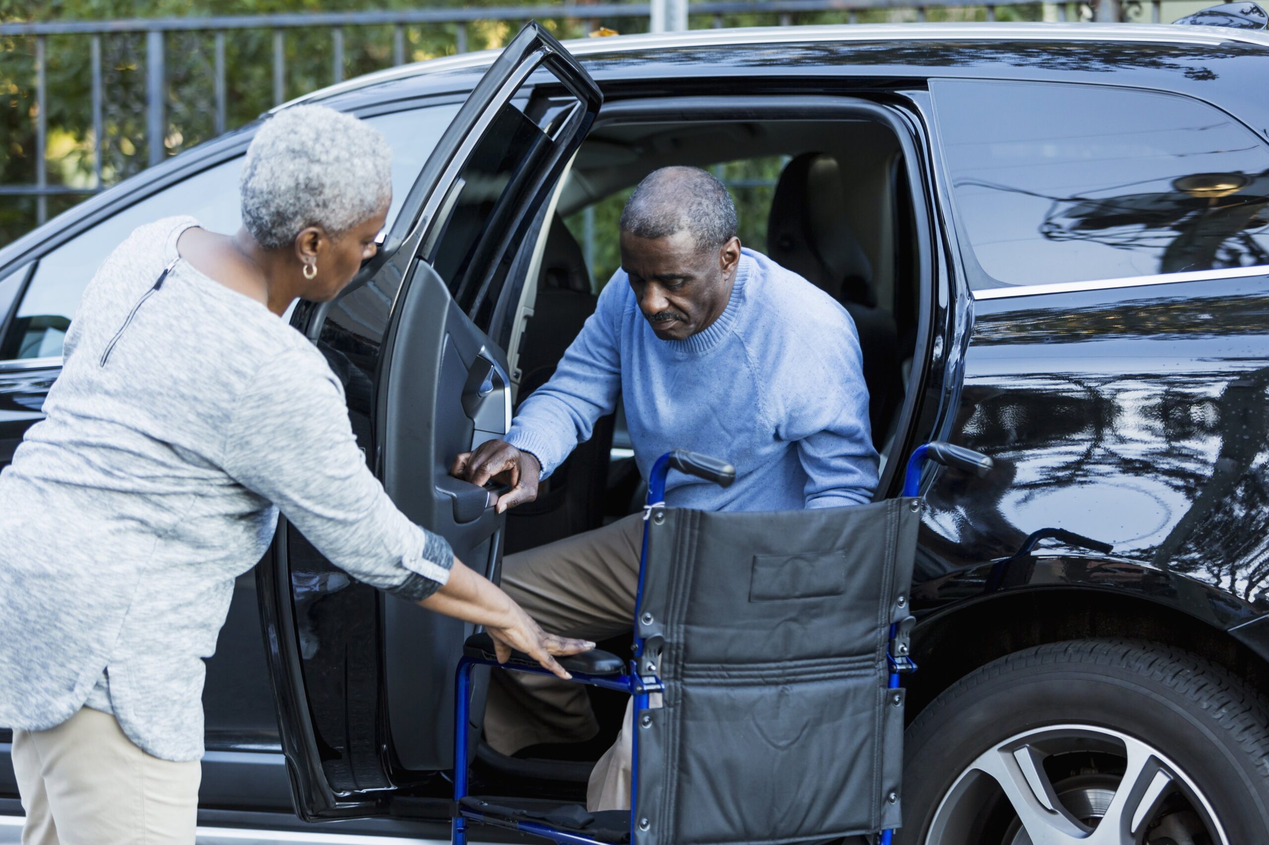 An elderly woman helping her husband from a car into a wheelchair