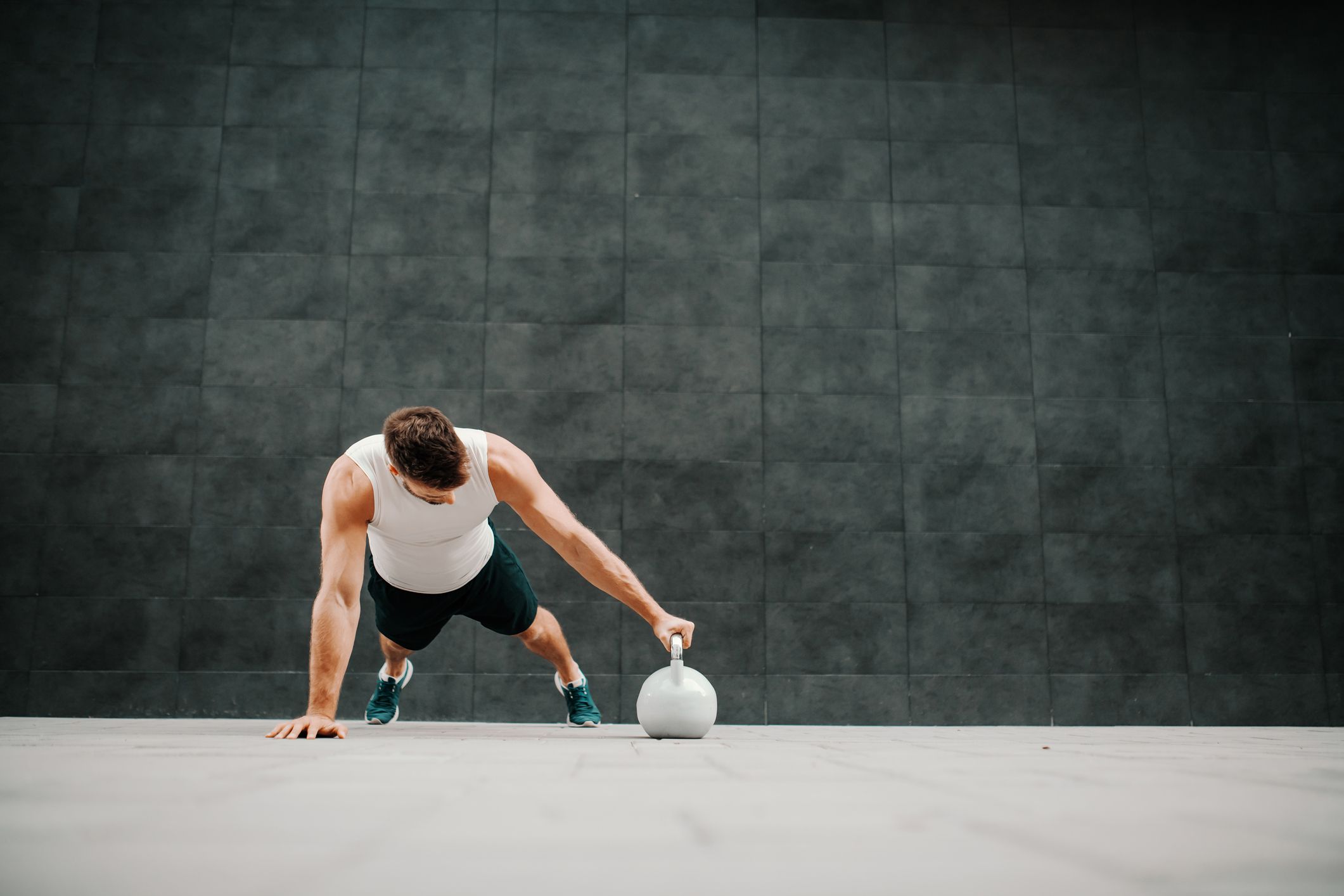 handsome muscular man in shorts and t-shirt doing push ups with kettle bell. In background is gray wall.