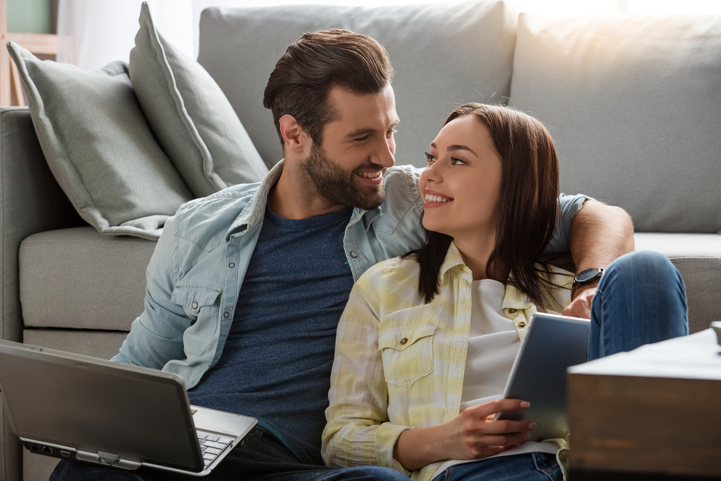 Couple using a laptop and tablet at home smiling at each other