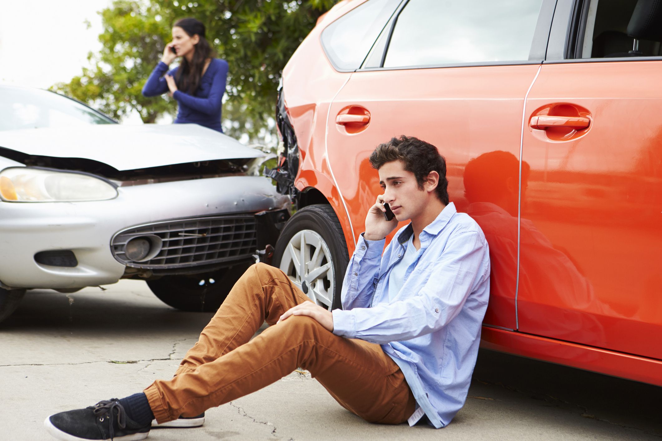 worried teen sitting on the pavement making phone call in front of car accident