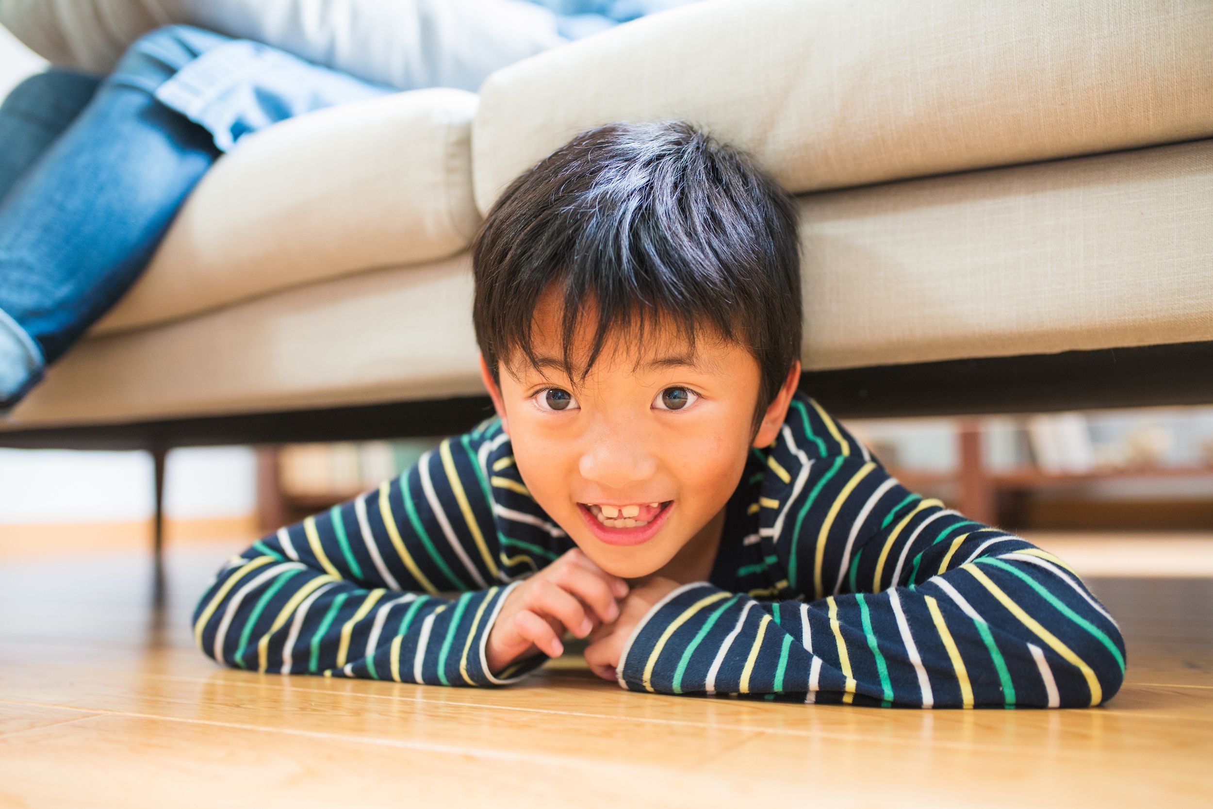 young boy with missing teeth, smiling while hiding under the sofa